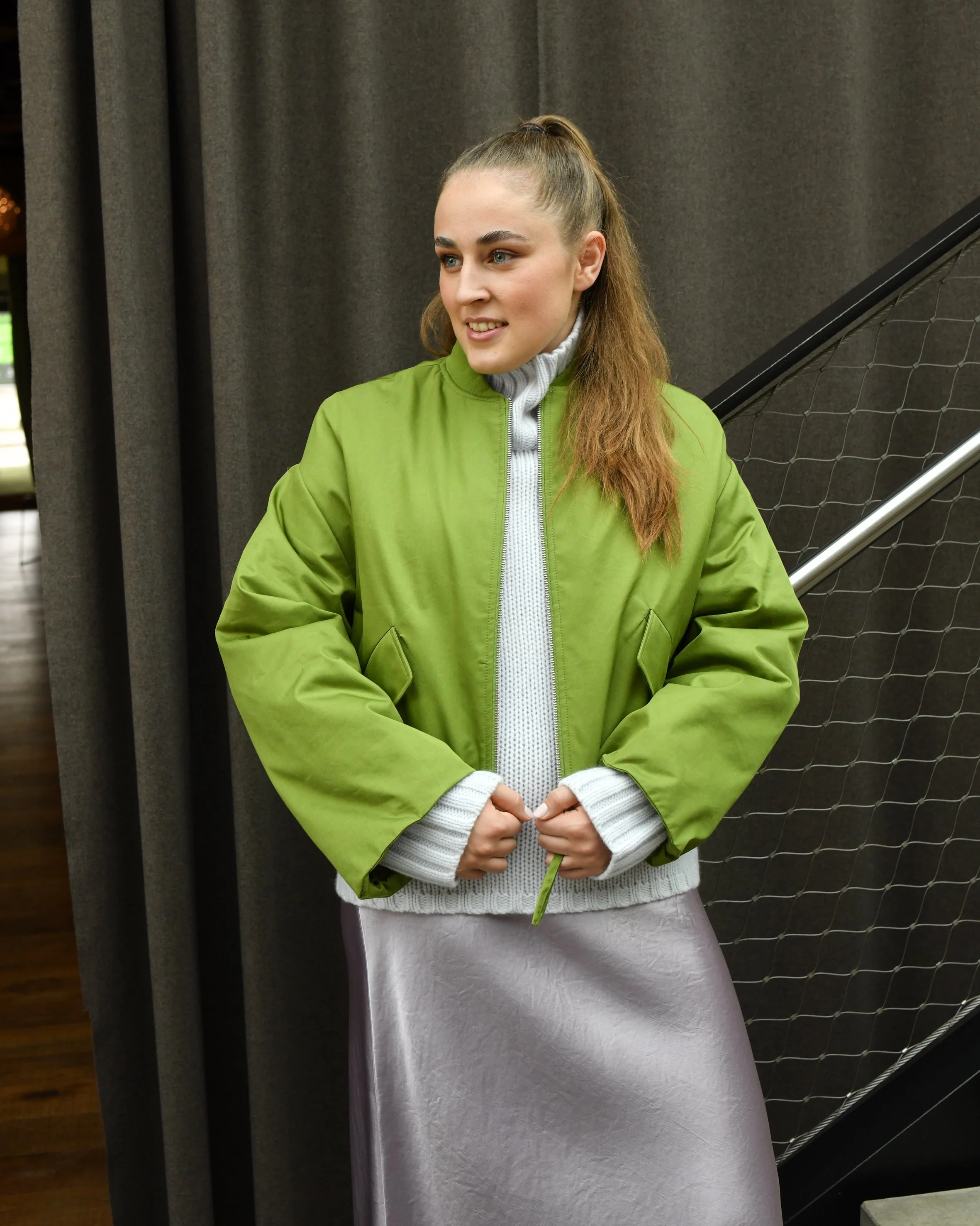 Frau mit braunen langen Haaren. Sie trägt einen Seidenrcok in rose, dazu einen grauen Rollkragenpullover und eine grüne Bomberjacke. 