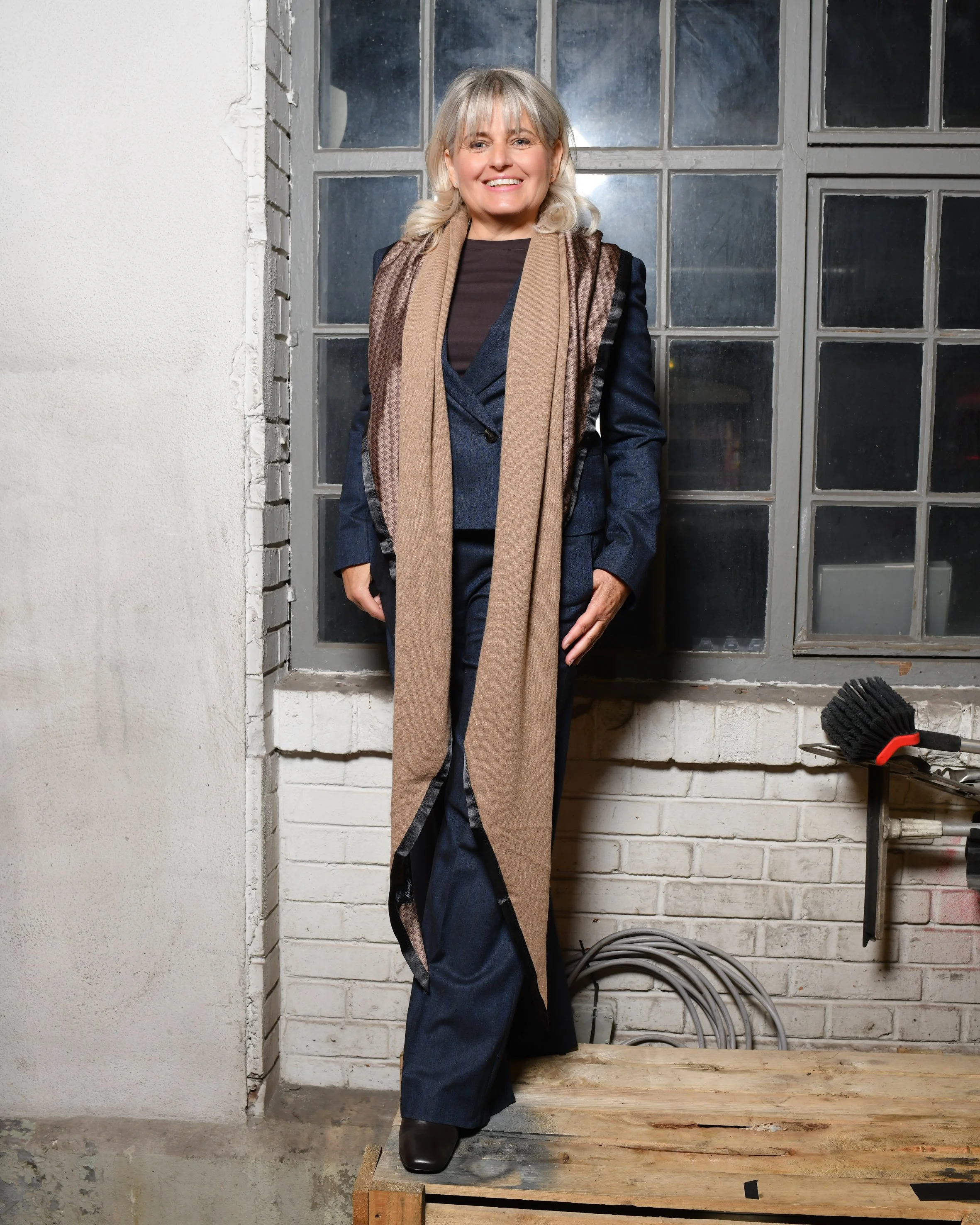 Frau trägt dunkelblauen, modernen Hosenanzug mit weiten Beinen und kurz geschnittenem Blazer. Dazu kombiniert einen hellbraunen langen Schal, eine silberne kleine Handtasche und schwarze elegante Schuhe.