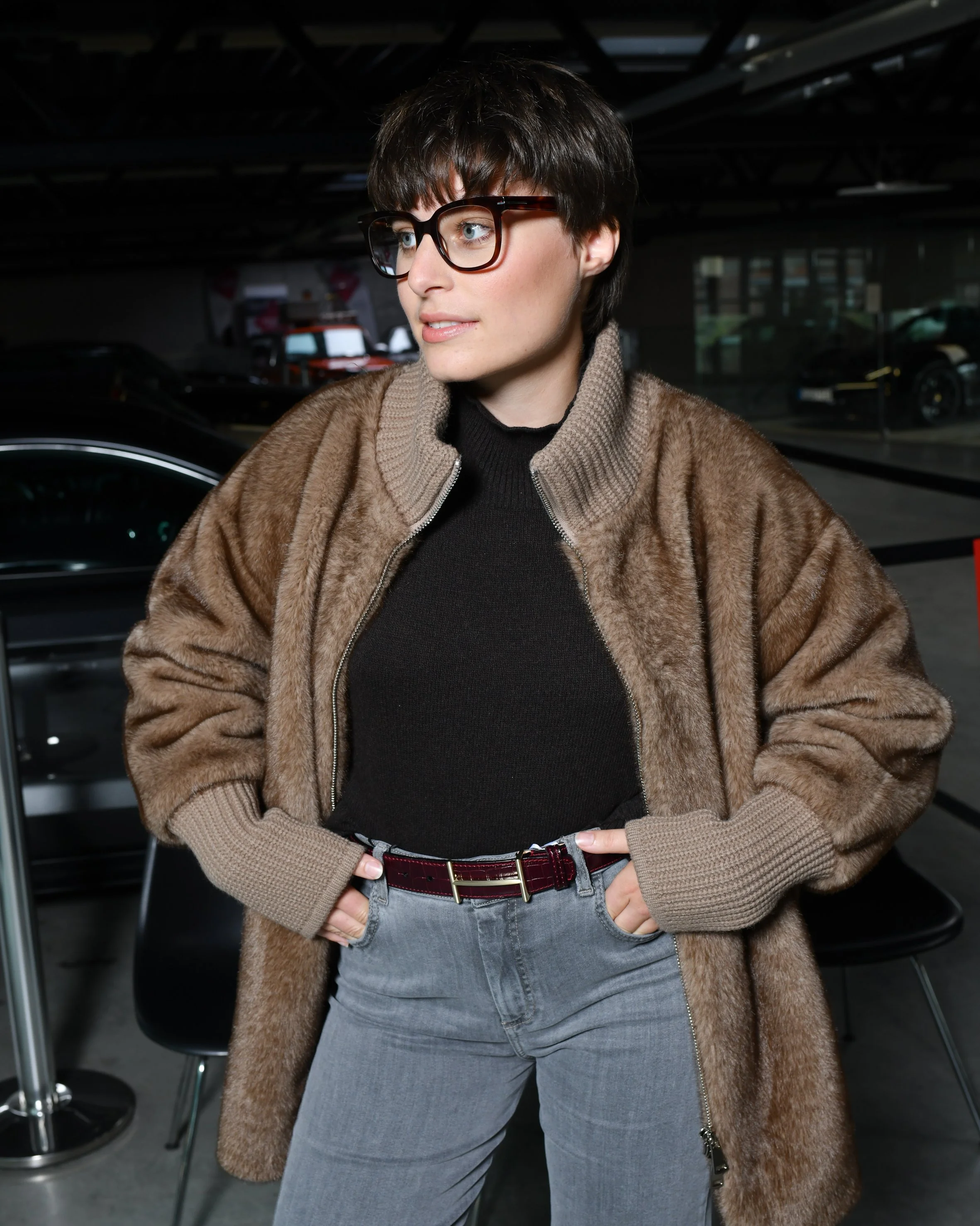 Frau mit grauer, weiter Jeans. Schwarzer Pullover und darüber eine braune kuschelige Jacke. Hohe Schuhe in braun. Sie steht und schaut nach links.