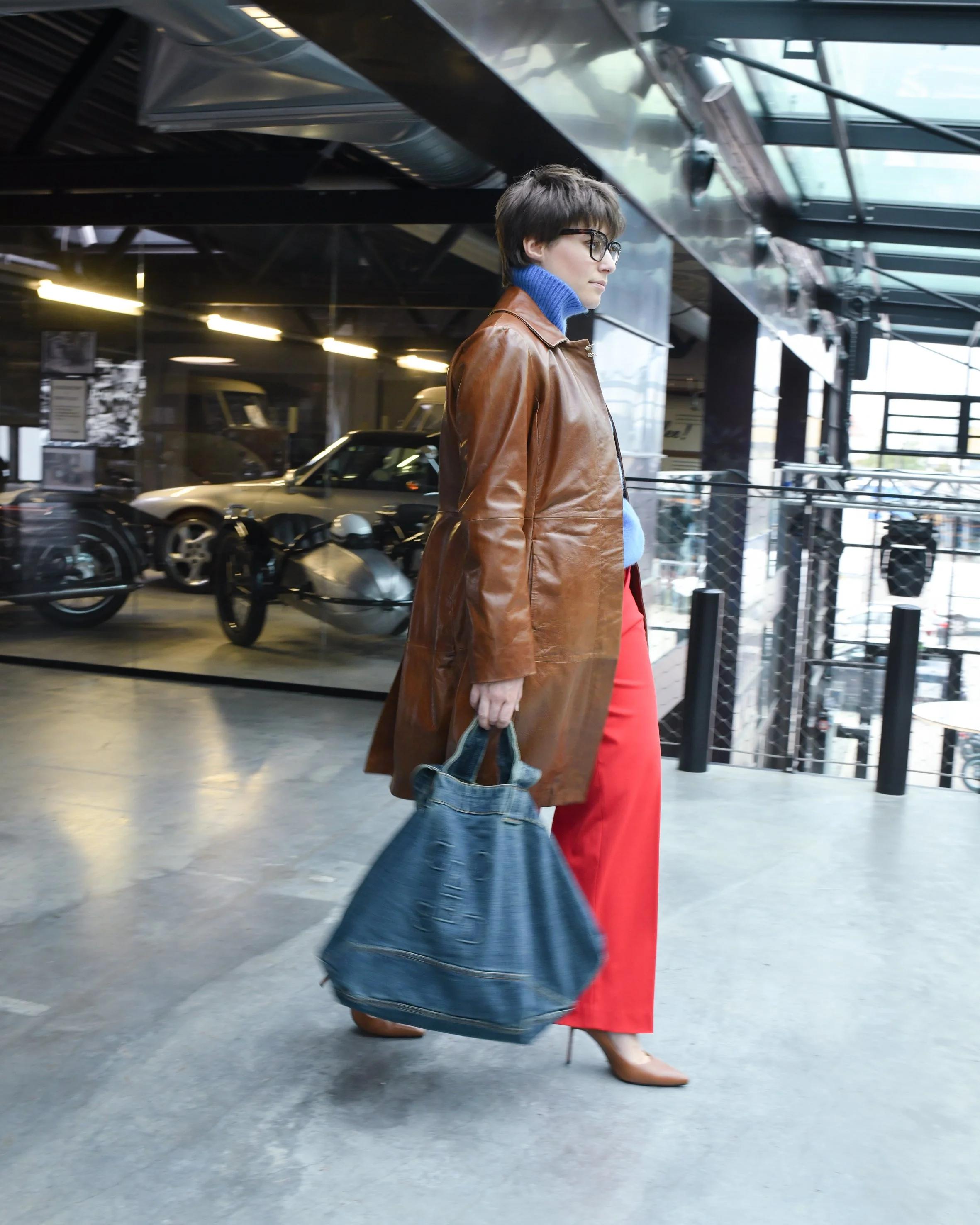 Frau mit kurzen braunen Haaren. Sie trägt eine lange rote Hose und einen blauen Rollkragenpullover. Darüber trägt sie einen langen braunen Ledermantel und braune Keilabsatzschuhe. In ihrer Hand hält sie eine Jeans Shopperbag von Closed.
