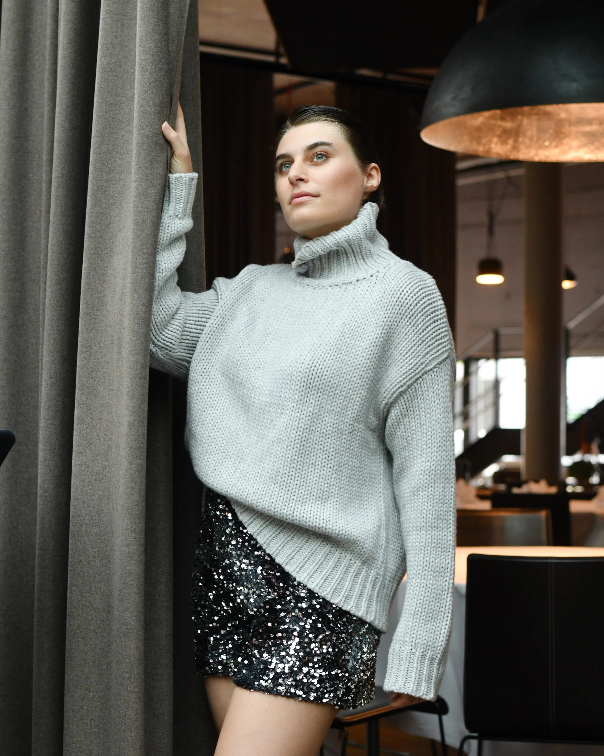 Eine Frau mit braunen kurzen Haaren. Sie trägt eine kurze schwarze Hose mit Pallietten und einen hellgrauen Rollkragenpullover.
