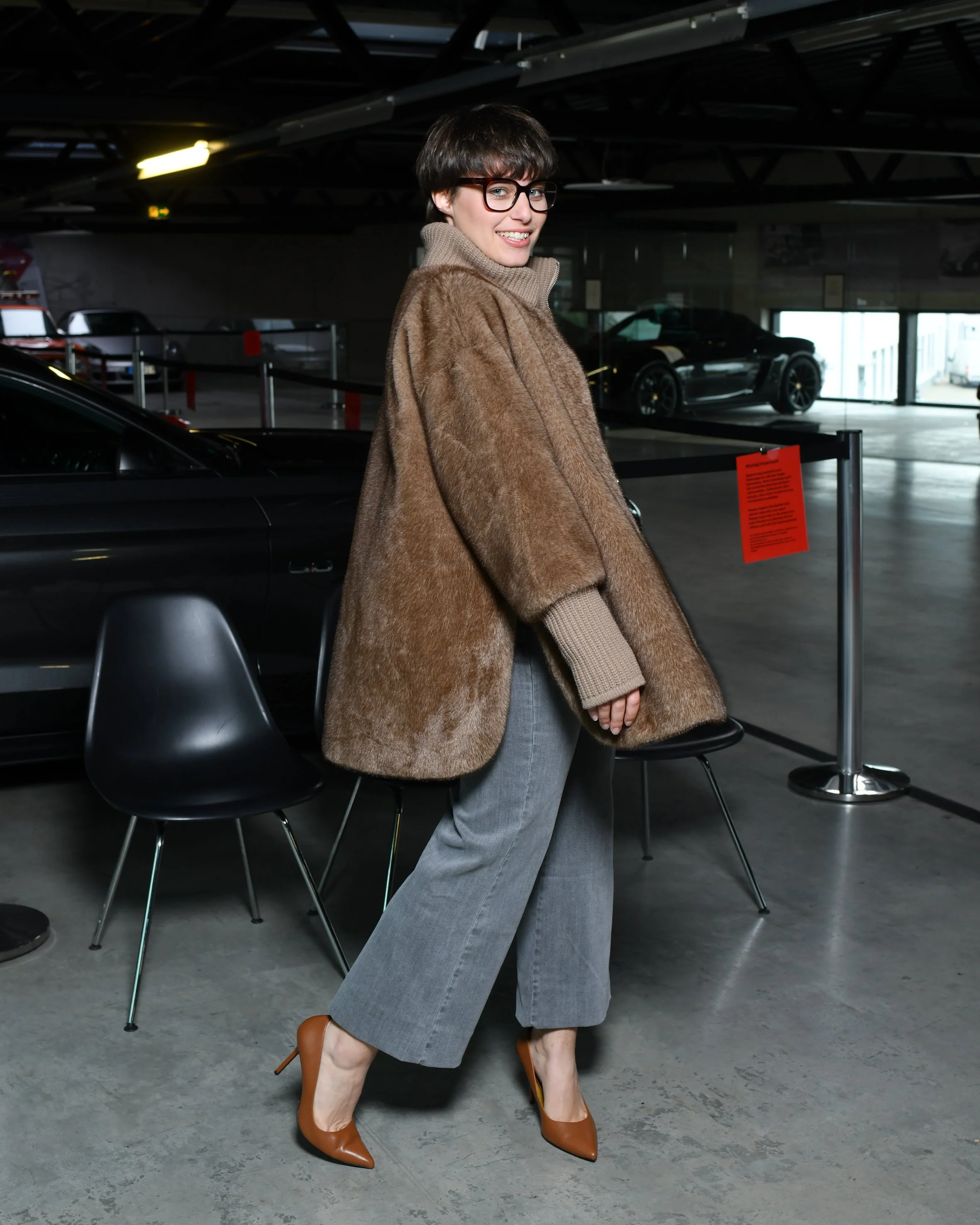 Frau mit grauer, weiter Jeans. Schwarzer Pullover und darüber eine braune kuschelige Jacke. Hohe Schuhe in braun. Sie steht und lächelt in die Kamera.