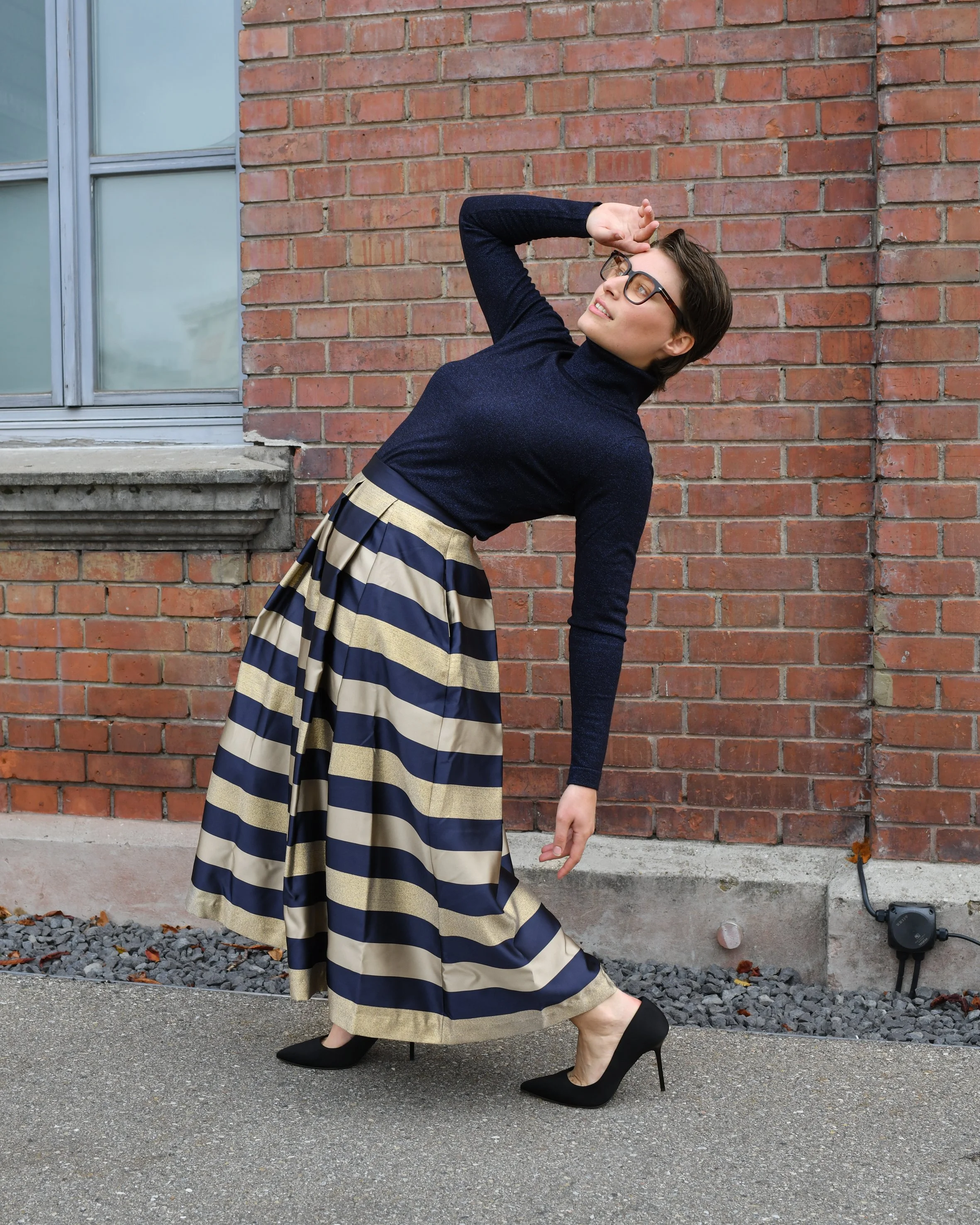 Eine Frau mit trägt einen langen gestreiften Rock in gold, dunkelblau. Dazu ein dunkelblaues Langarmshirt und schwarze elegante Absatzschuhe.