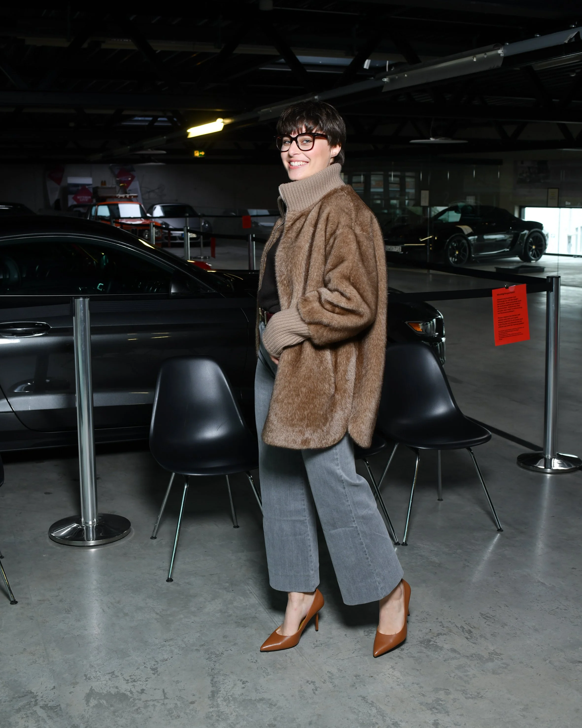 Frau mit grauer, weiter Jeans. Schwarzer Pullover und darüber eine braune kuschelige Jacke. Hohe Schuhe in braun. Sie steht und lächelt in die Kamera.