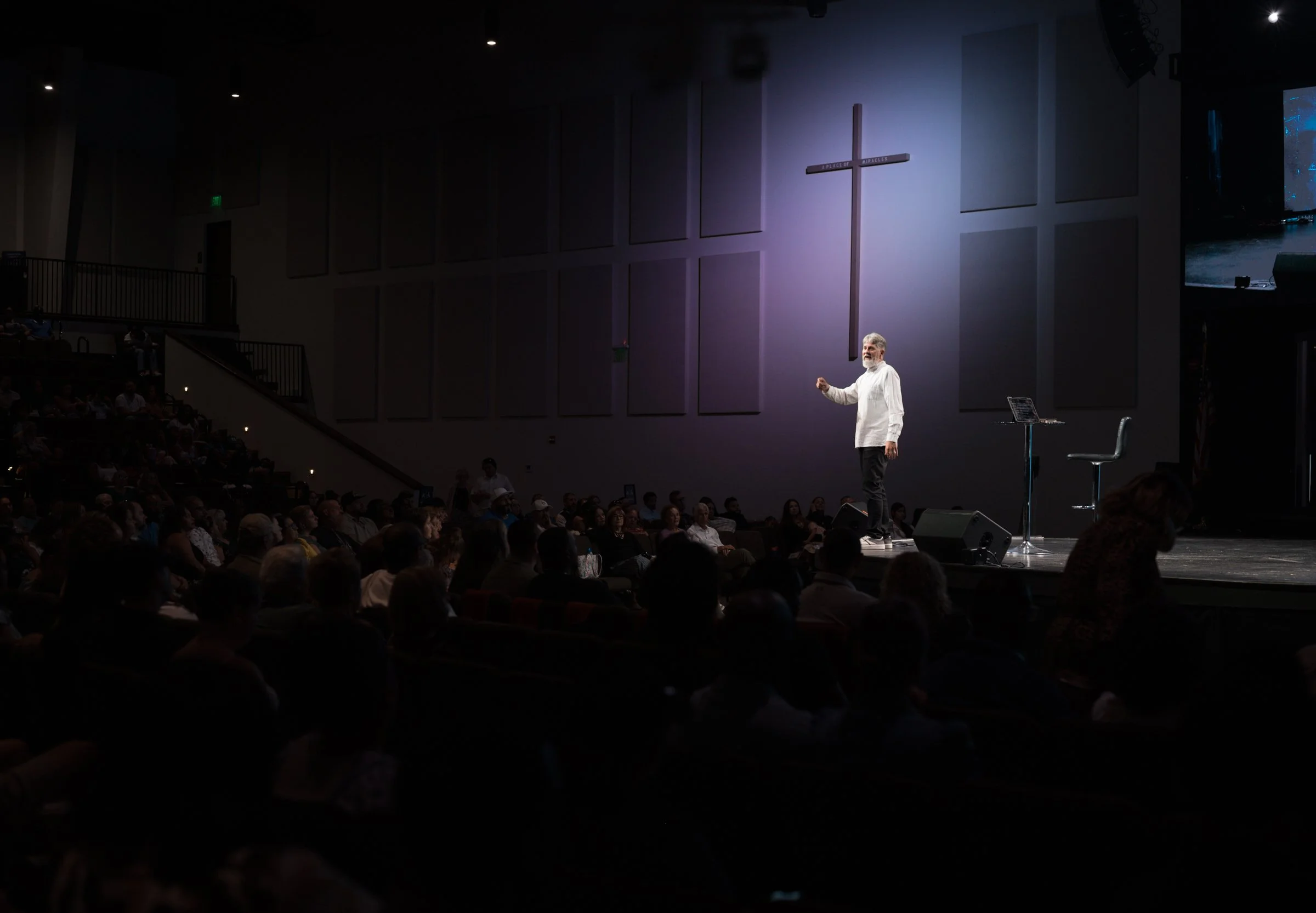 A speaker standing on stage in front of a church cross, addressing an audience in a dark auditorium.