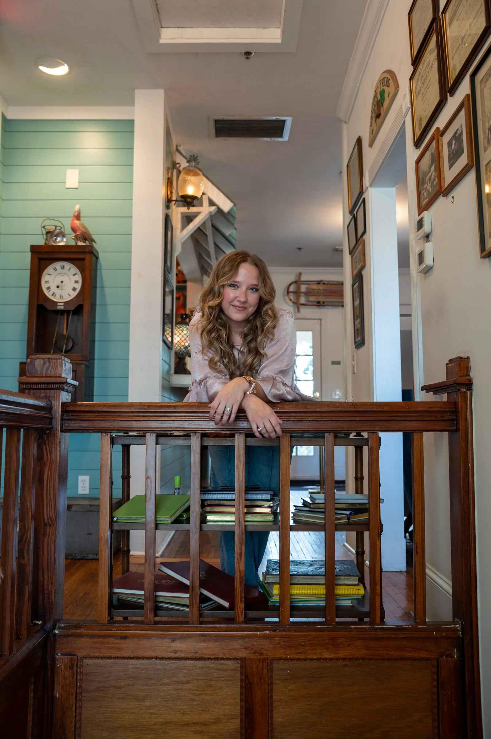 A woman with long curly blonde hair leaning on a wooden railing inside a cozy home, smiling at the camera, with books on the lower shelf of the railing, framed pictures on the wall, a clock, and decorative items visible in the background.