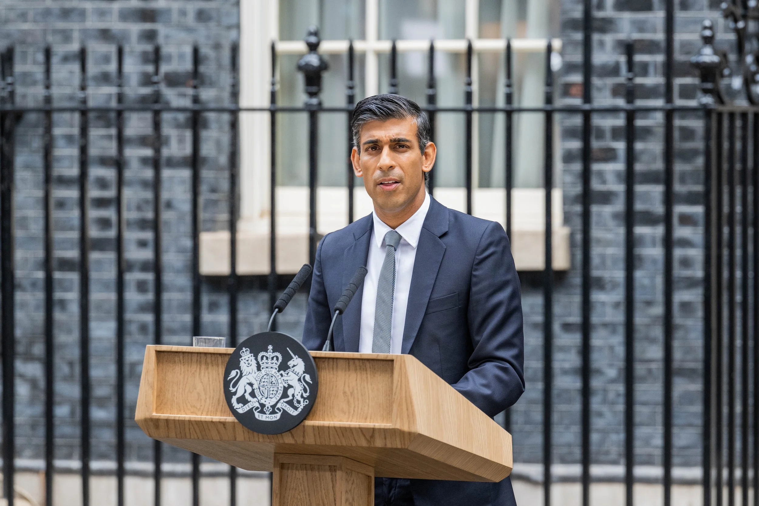 A man in a dark suit standing behind a wooden podium with a government emblem, speaking outdoors in front of a black iron fence and grey brick building.
