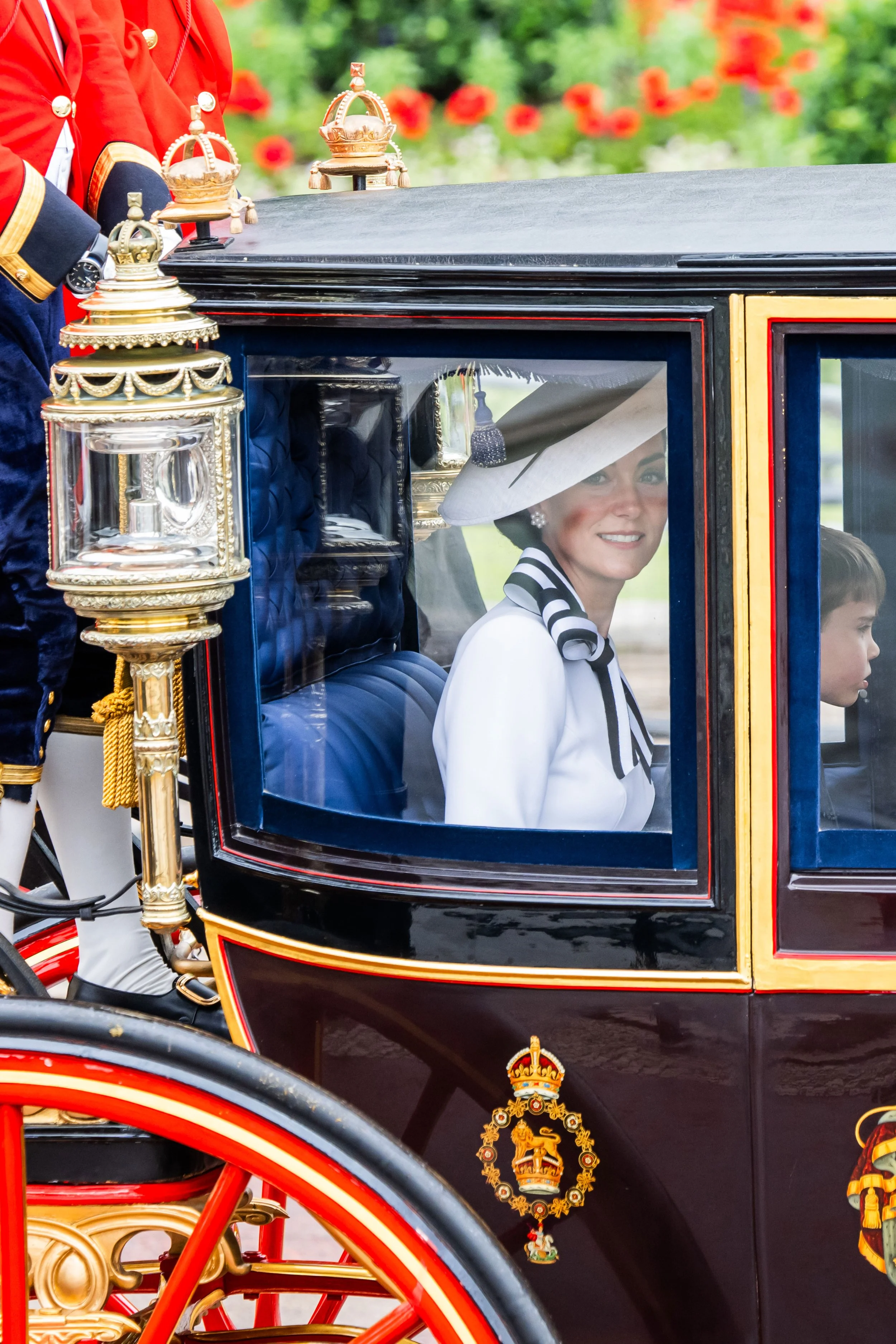 A woman dressed in white with a large hat and black-and-white scarf sitting inside a royal carriage, smiling, during a formal event or procession.
