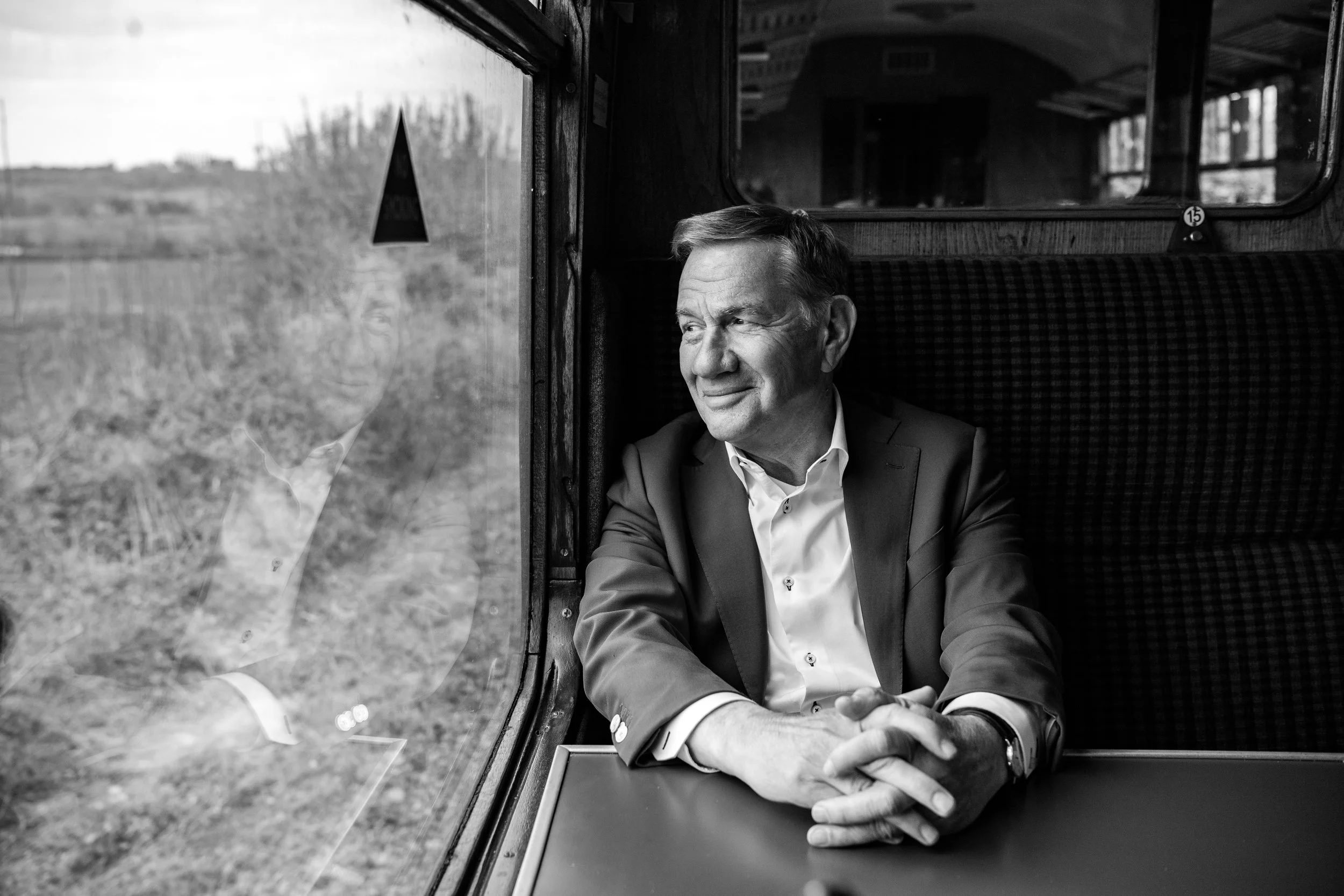 A man in a suit sitting at a train window, looking outside thoughtfully with his hands clasped on the table.