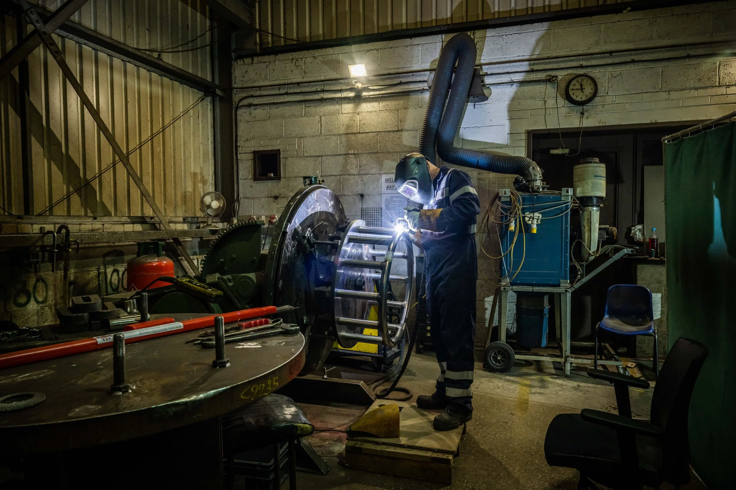 A worker wearing protective gear welding metal inside an industrial workshop with tools, machinery, and a clock on the wall.