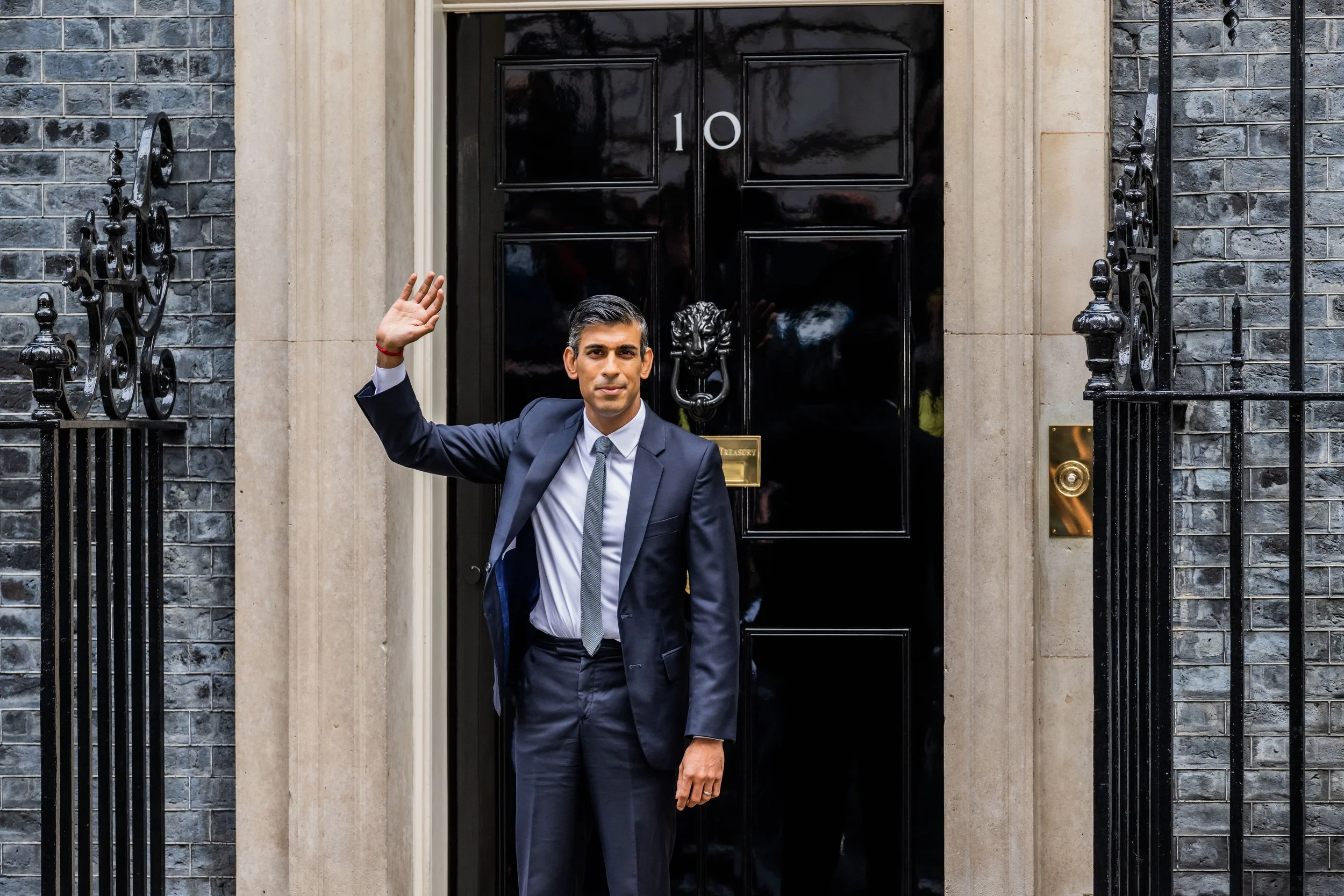 A man in a suit standing in front of a black door with a lion head door knocker, waving with his right hand.