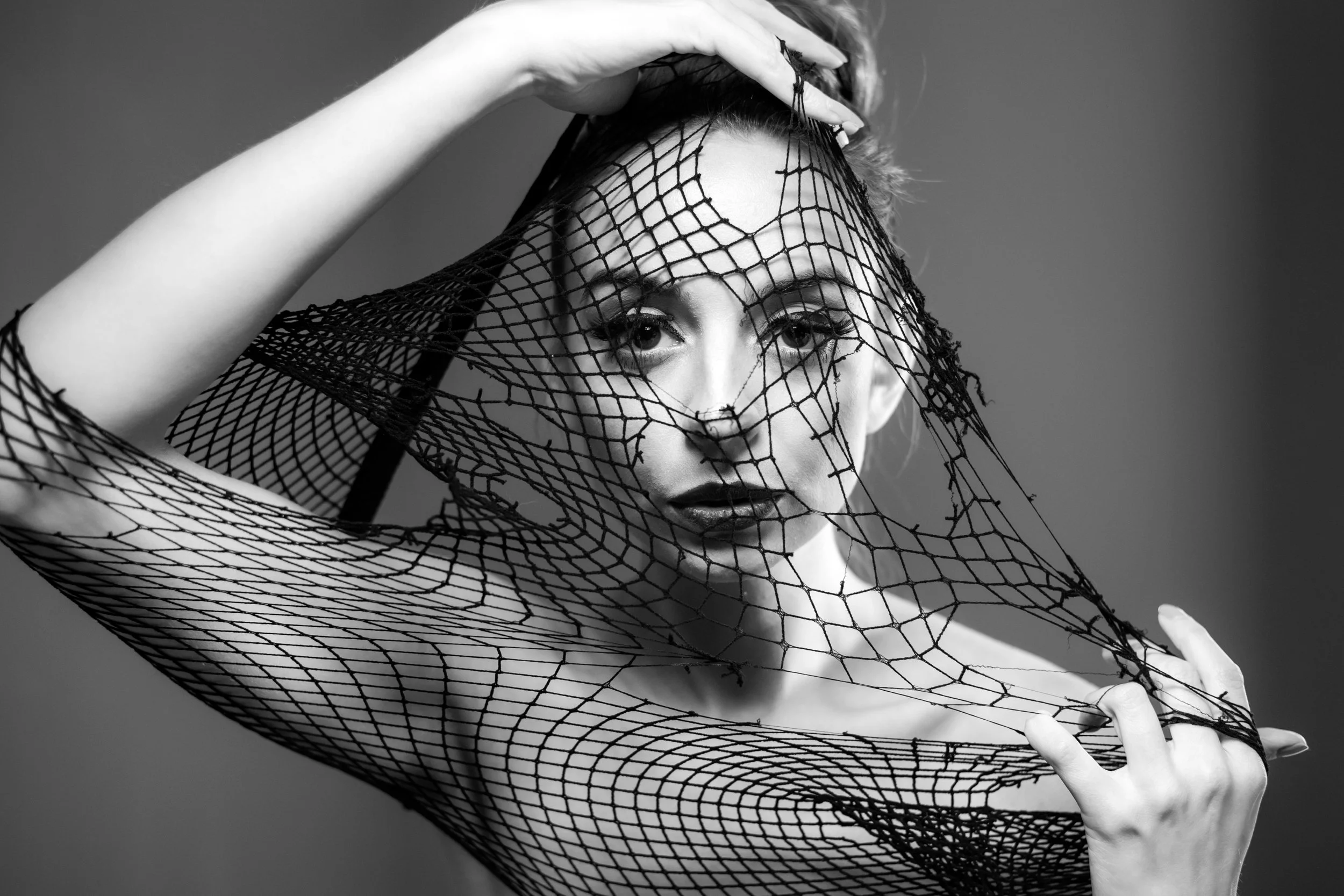 A black and white portrait of a woman with a fishnet veil covering her face and arm, striking a pose with her hand on her head and shoulder.