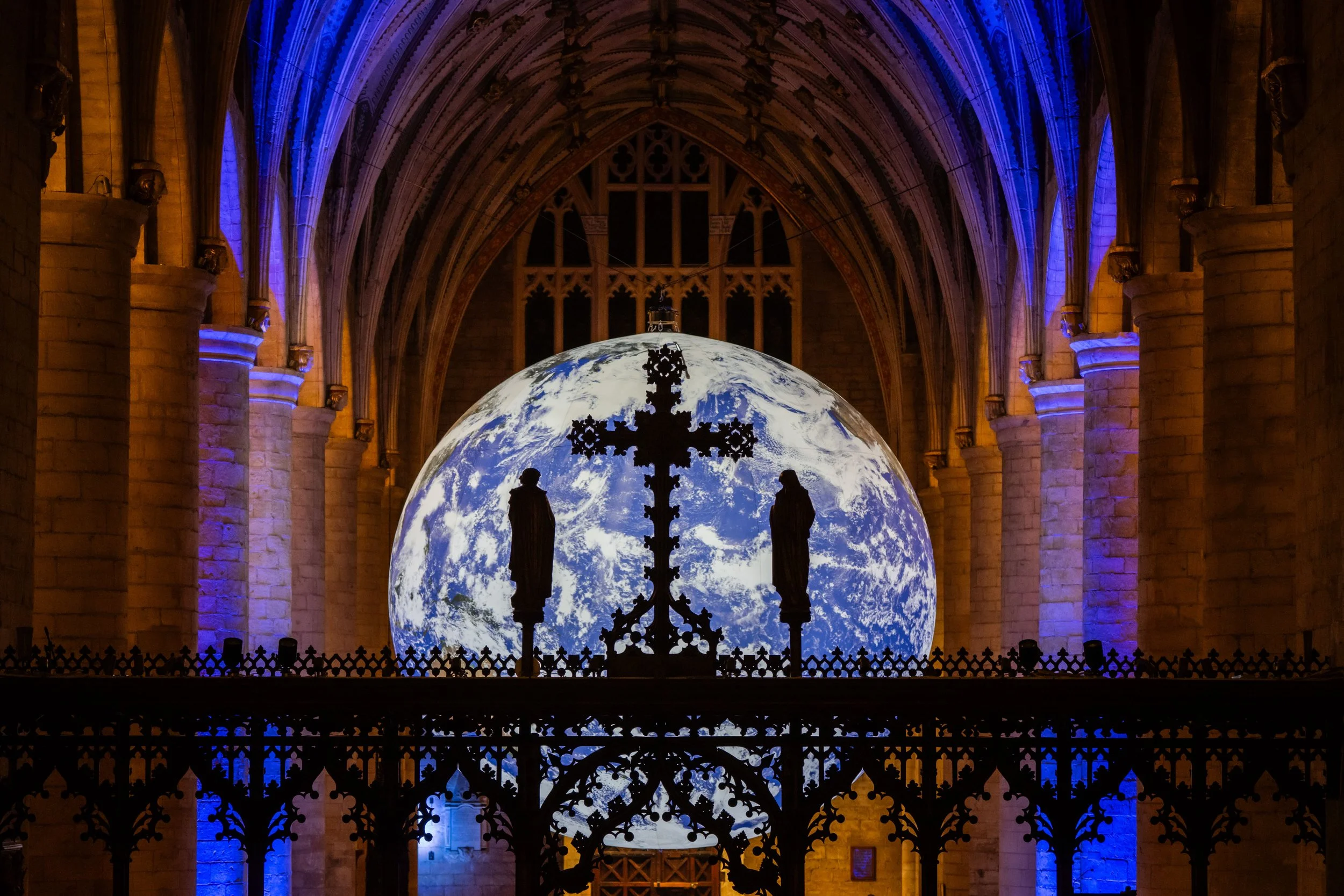 Inside a cathedral with stained glass and arches, a large globe of Earth is displayed in the background, illuminated with a blue hue. Silhouettes of two statues and an ornate cross are in the foreground.