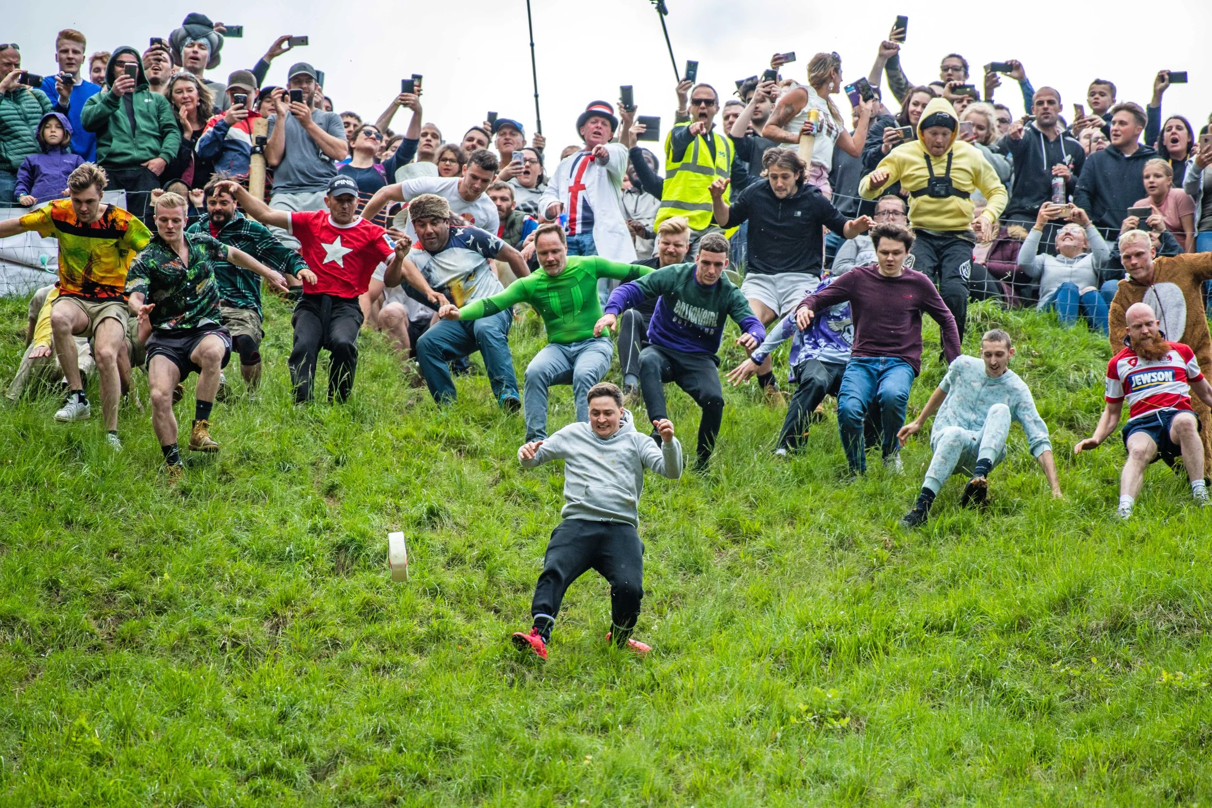 People participating in a grass hill race, with spectators watching and recording from above.