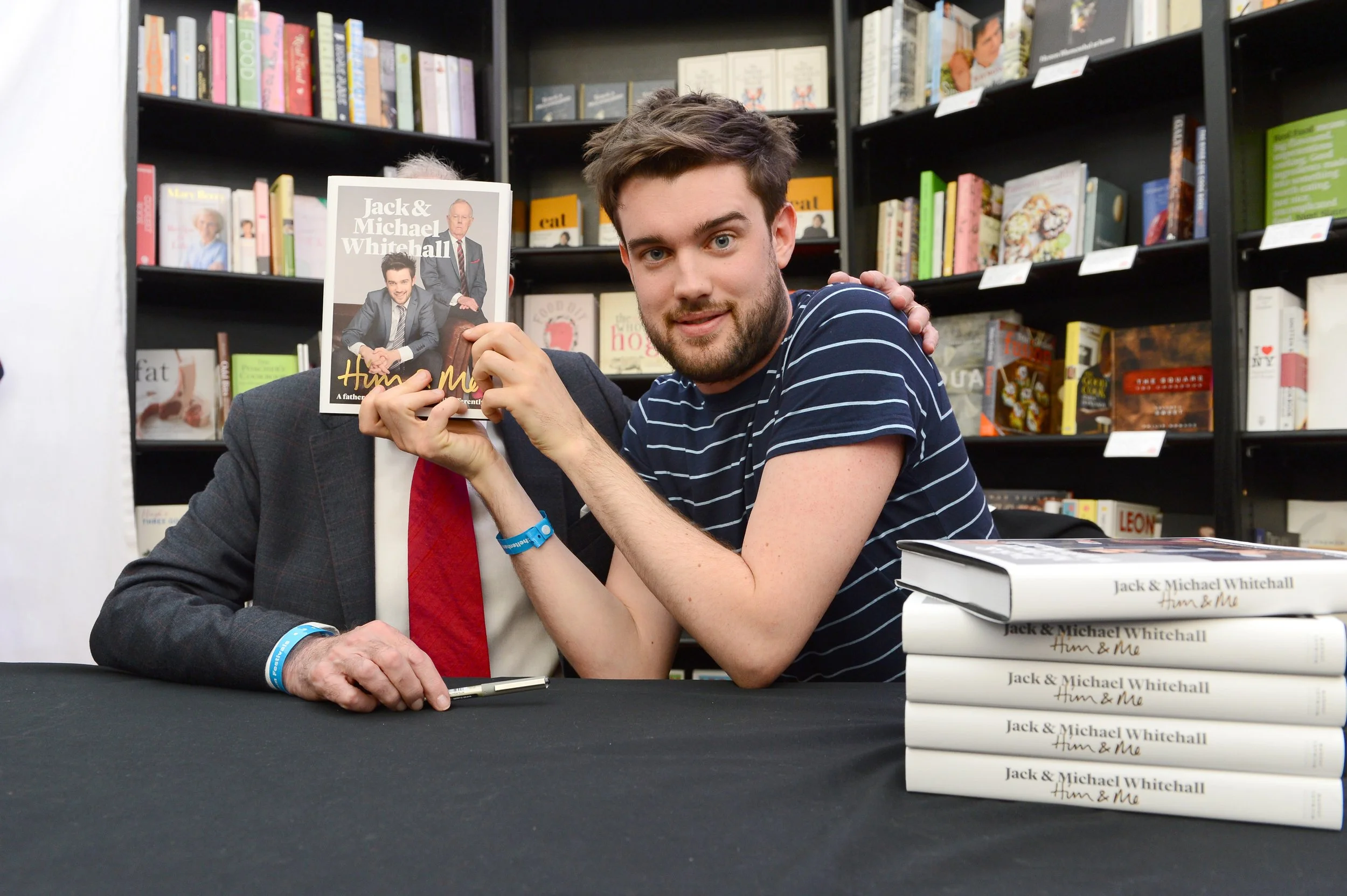 A young man with a beard and striped shirt holding a book in a bookstore or library, with bookshelves behind him and a stack of books on the table in front of him.