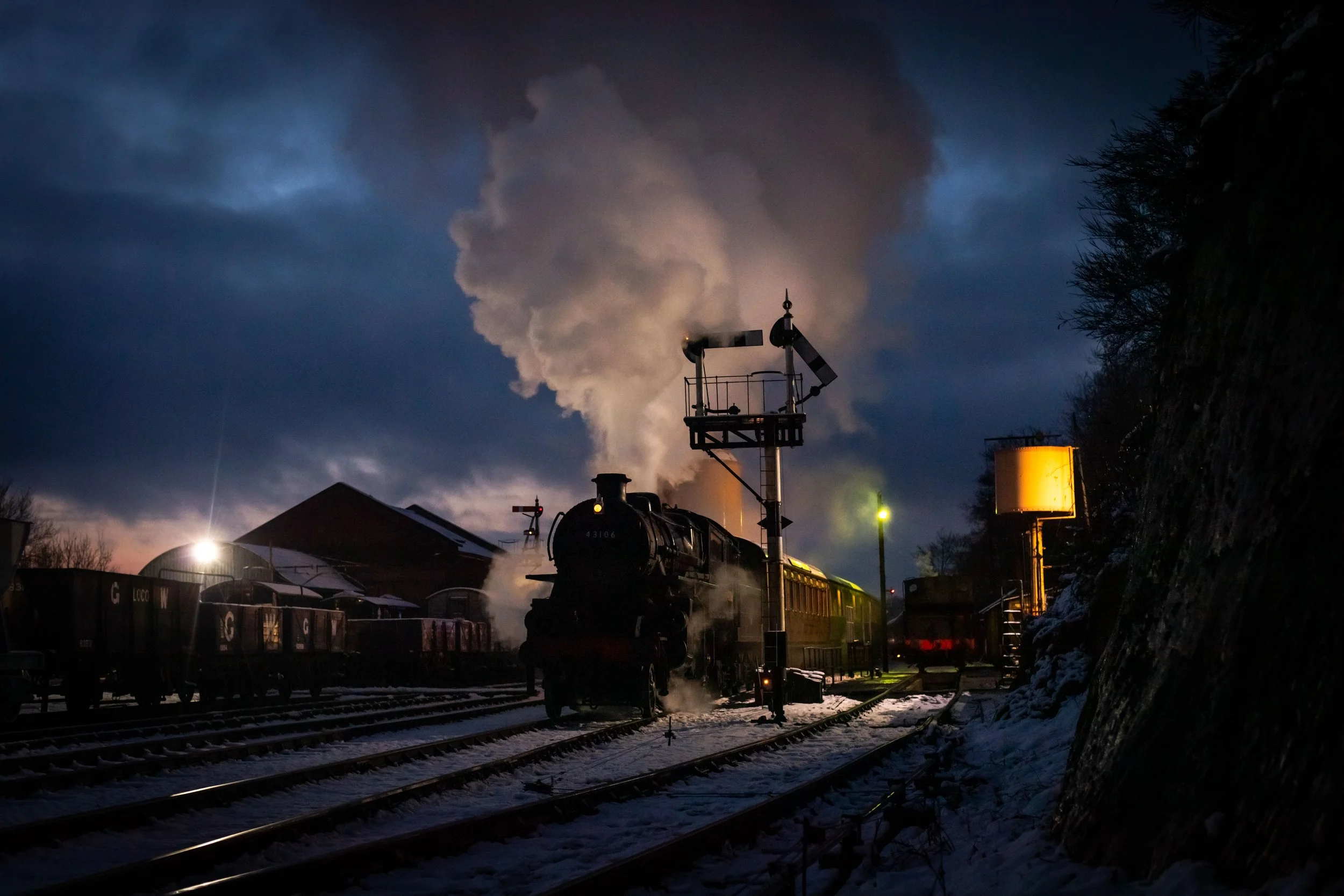 A steam train on snow-covered tracks at dusk or night, emitting steam and smoke, with station signals and buildings in the background.