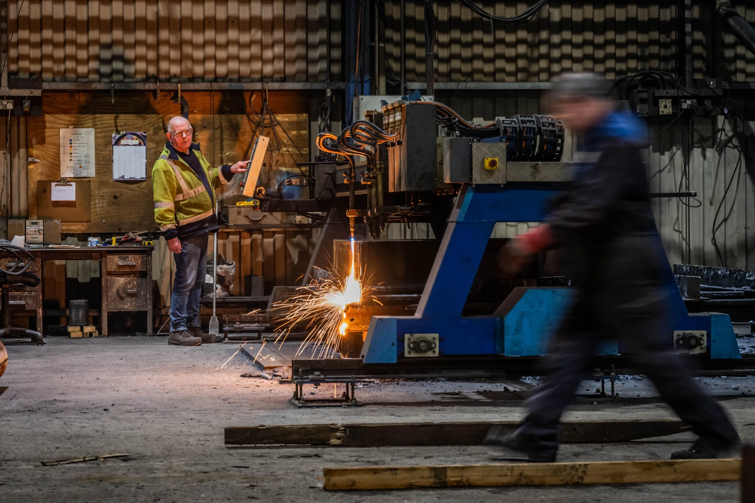 Two workers in a workshop, one standing, observing, and the other walking past a large blue industrial machine, with sparks flying as the machine operates.