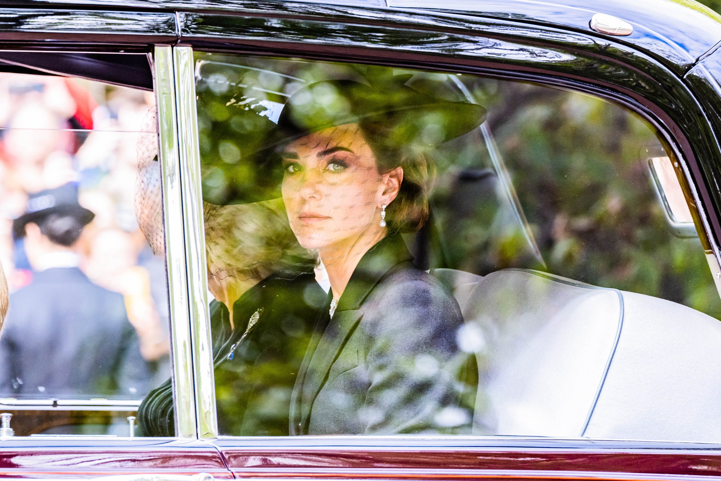 A woman dressed in vintage attire sitting inside a car, seen through the window with reflections of greenery and blurred figures outside.