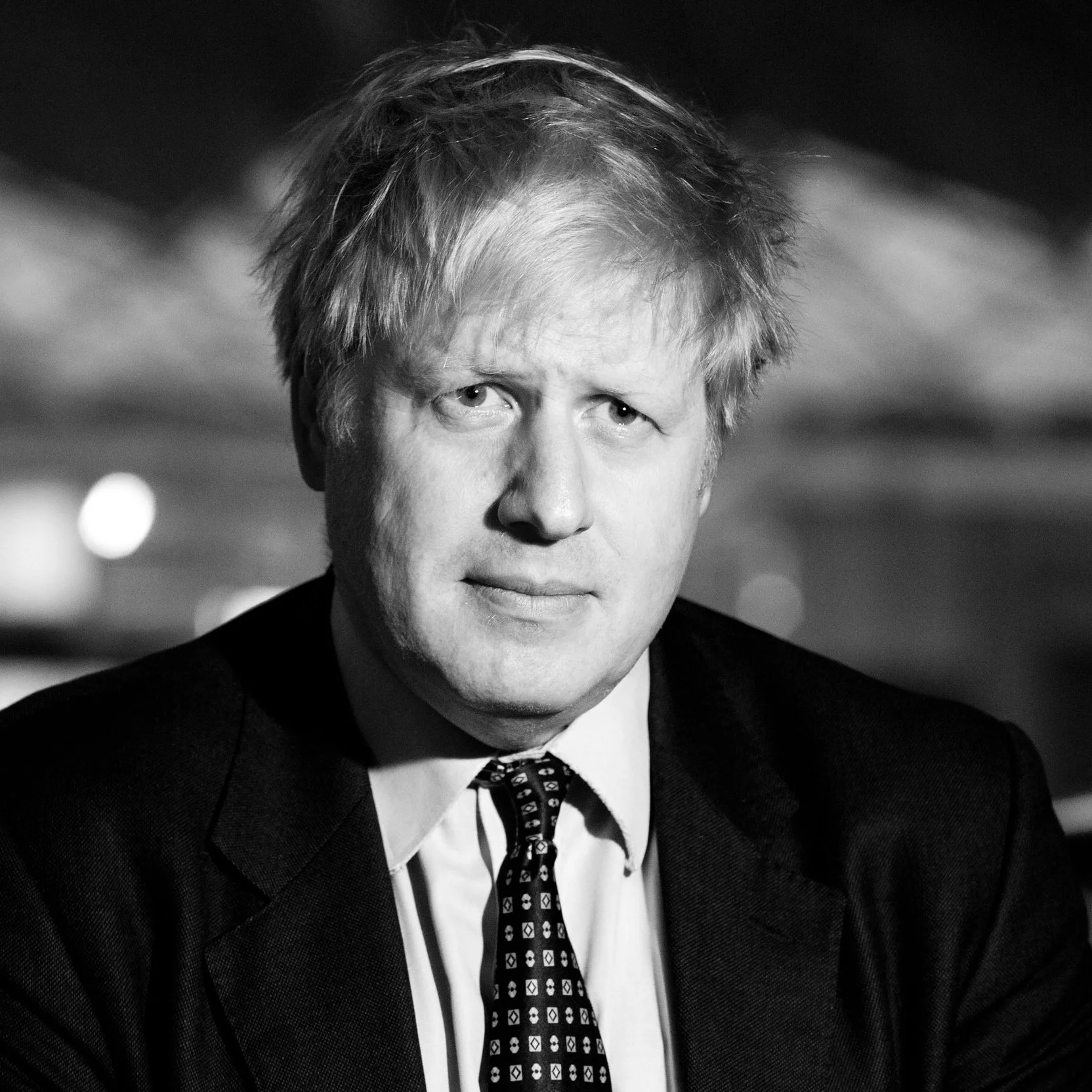 A black and white photograph of a man with messy hair wearing a suit, white shirt, and patterned tie, outdoors at night.