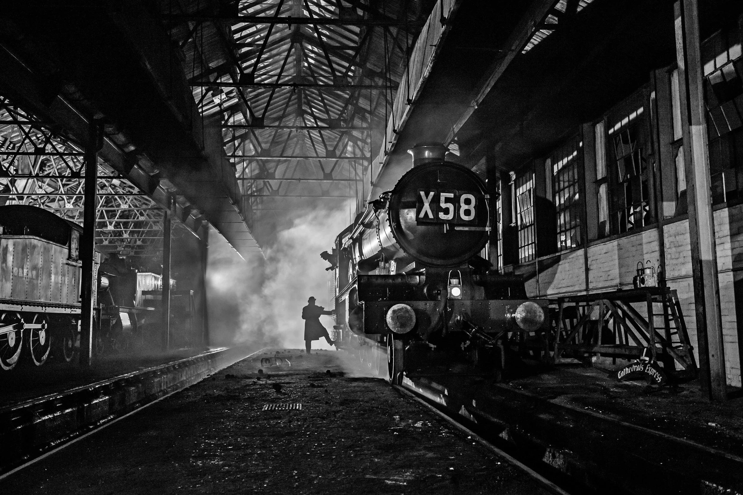 A vintage steam locomotive inside a train shed, with a person working near the front of the train, with smoke and steam visible in the air, black and white photo.