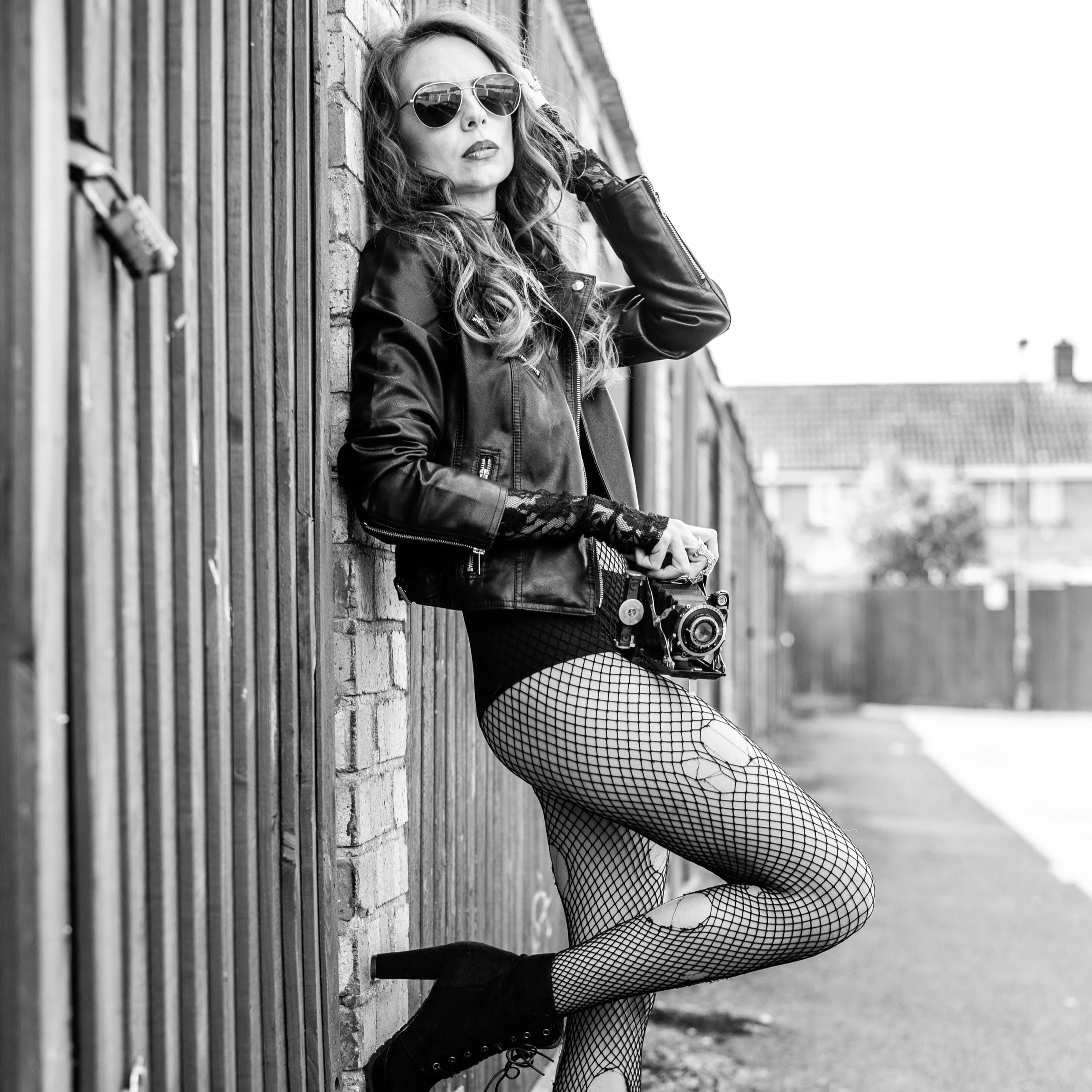 A woman posing against a brick wall, wearing sunglasses, a leather jacket, fishnet stockings, and black shoes, holding a camera.