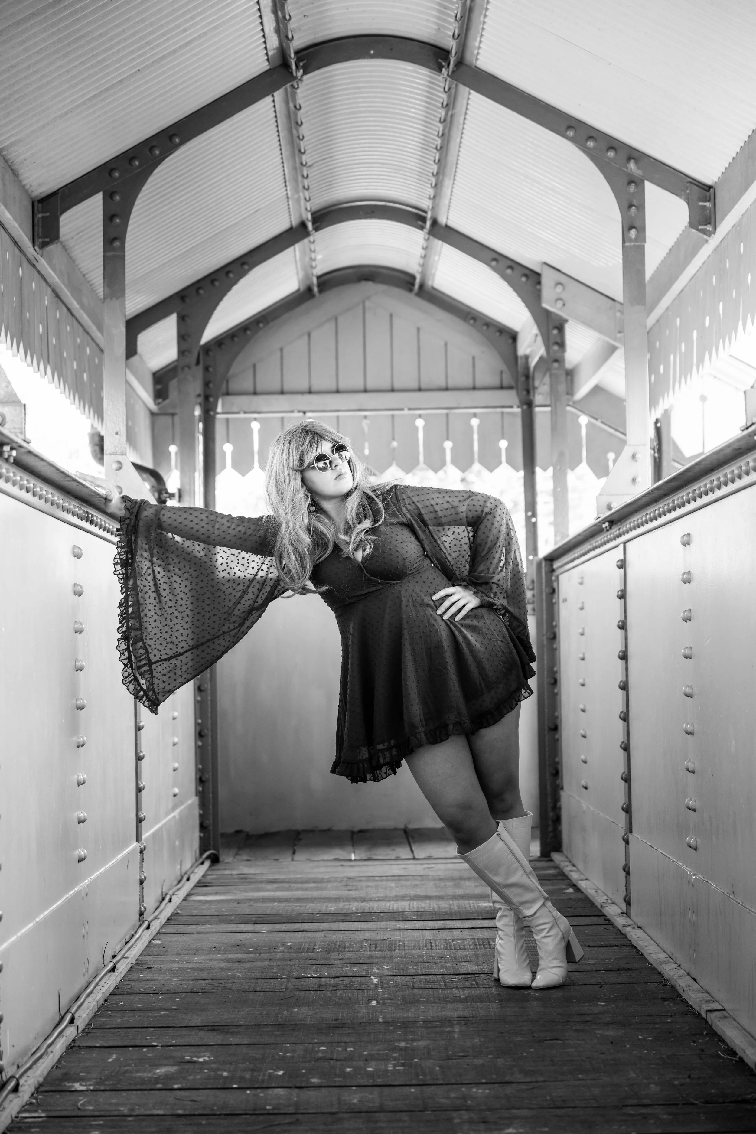 A woman in a black dress and white boots posing inside a covered walkway, wearing sunglasses and looking away.