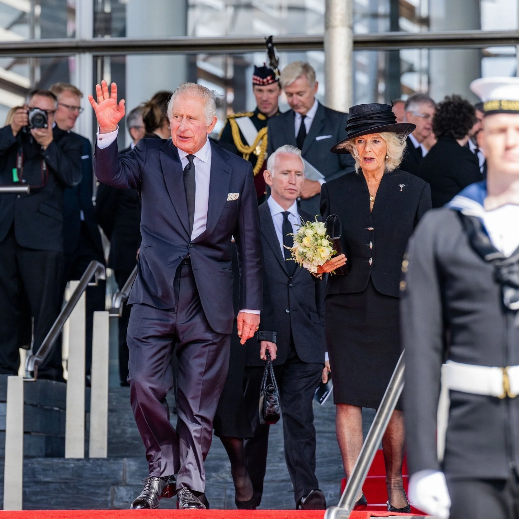 An elderly man in a navy suit and white shirt waves as he walks down the steps of a building. A woman in a black dress and hat holds a bouquet of flowers as they descend. Several other formally dressed individuals and a photographer are visible in the background.