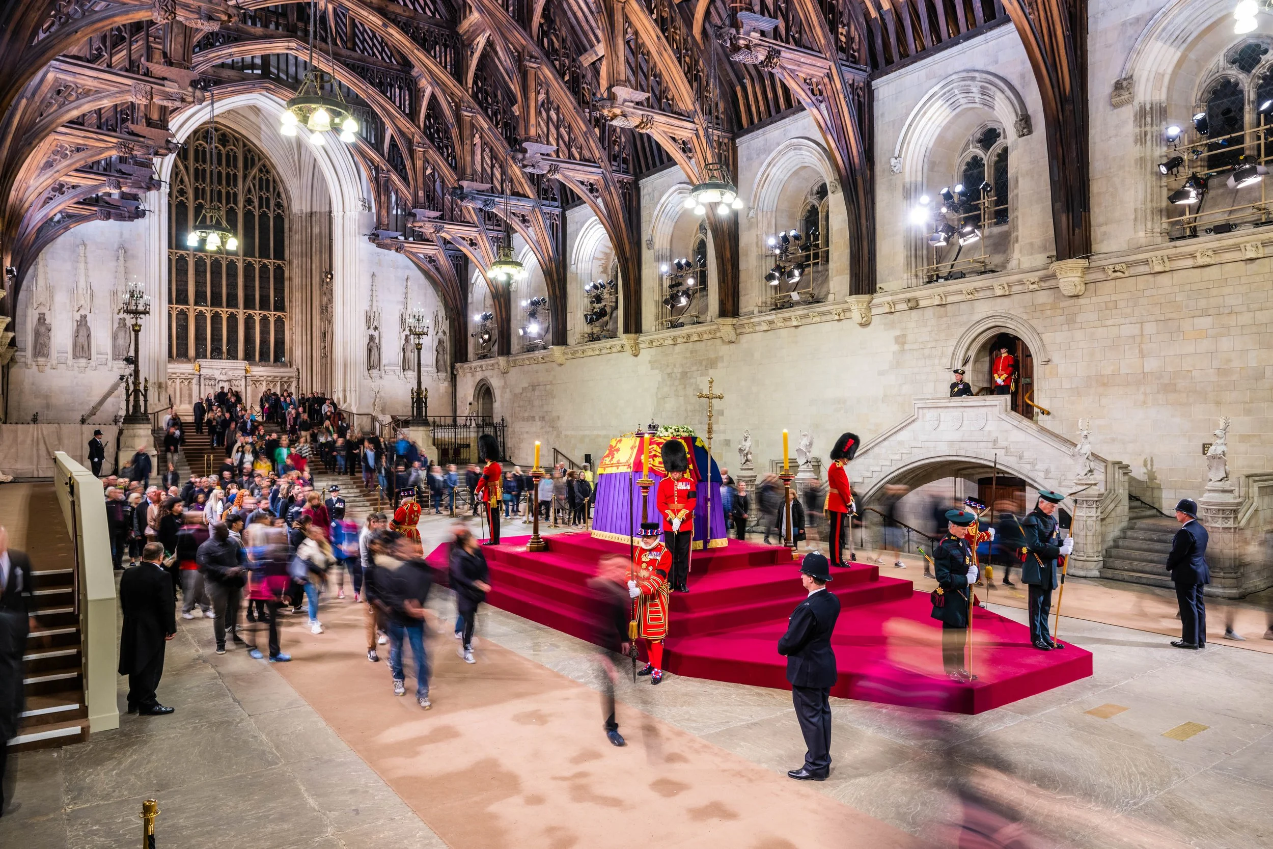 Interior of Westminster Abbey during the funeral of Queen Elizabeth II, with a platform containing the coffin draped in a purple and gold cloth, surrounded by guards in red and black ceremonial uniforms, and visitors walking through the church.