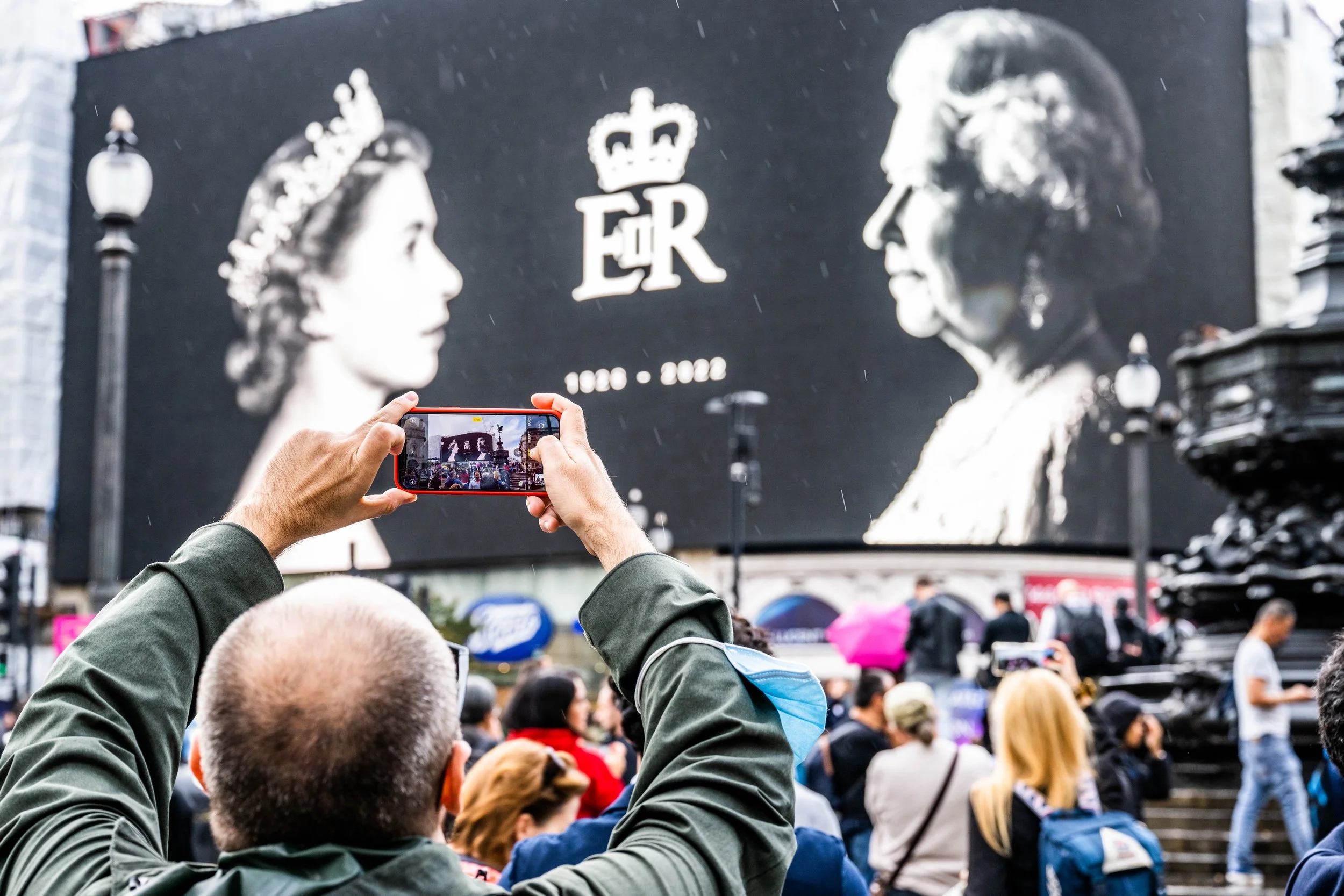 Crowd of people gathered outdoors, taking photos of a large mural depicting Queen Elizabeth II and a royal crown, with the text "E R" and the years 1926-2022, near a fountain and street lamps. The scene appears rainy with some people holding umbrellas.