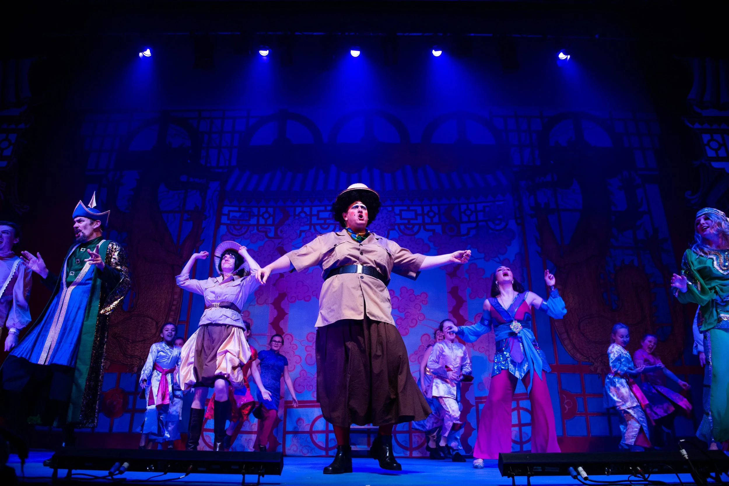 A theatrical stage performance with actors in colorful costumes, under spotlights, with a detailed set background featuring traditional Asian architectural elements.