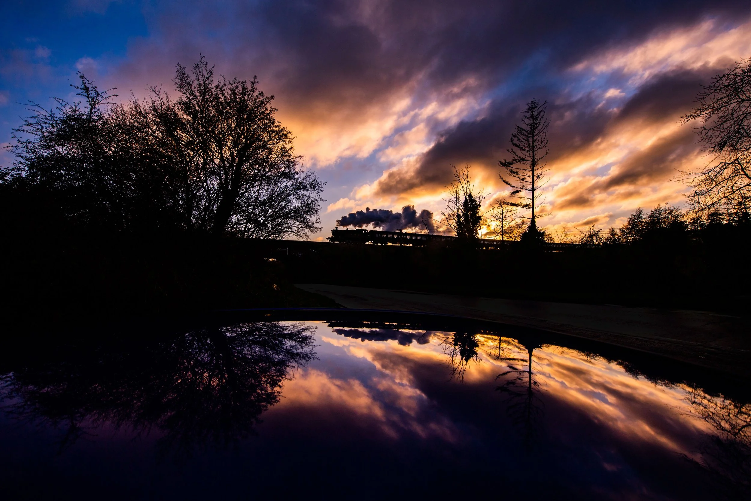 A sunset sky with colorful clouds, silhouettes of trees, a train emitting smoke, and a reflection of the sky and trees on a water surface.
