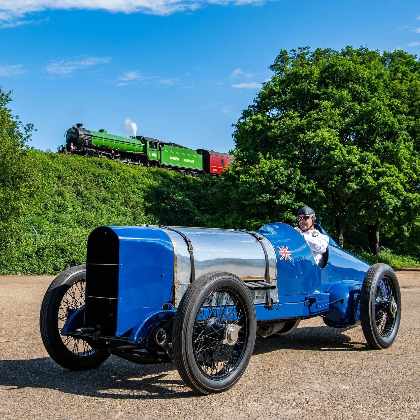A vintage blue race car with a UK flag decal on the side, driven by a person wearing goggles and a white jacket, parked on a paved area. In the background, a green and red steam locomotive labeled 'British Railways' is on a hill among green trees, under a clear blue sky.