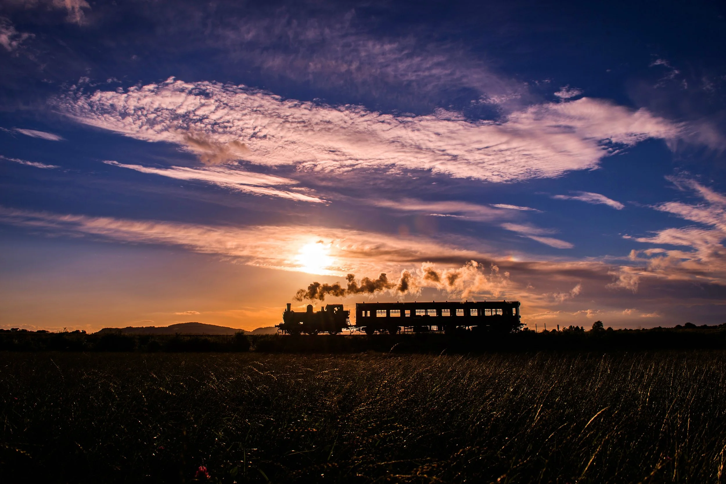 Silhouette of a moving steam train on a track during sunset with a colorful sky and scattered clouds.
