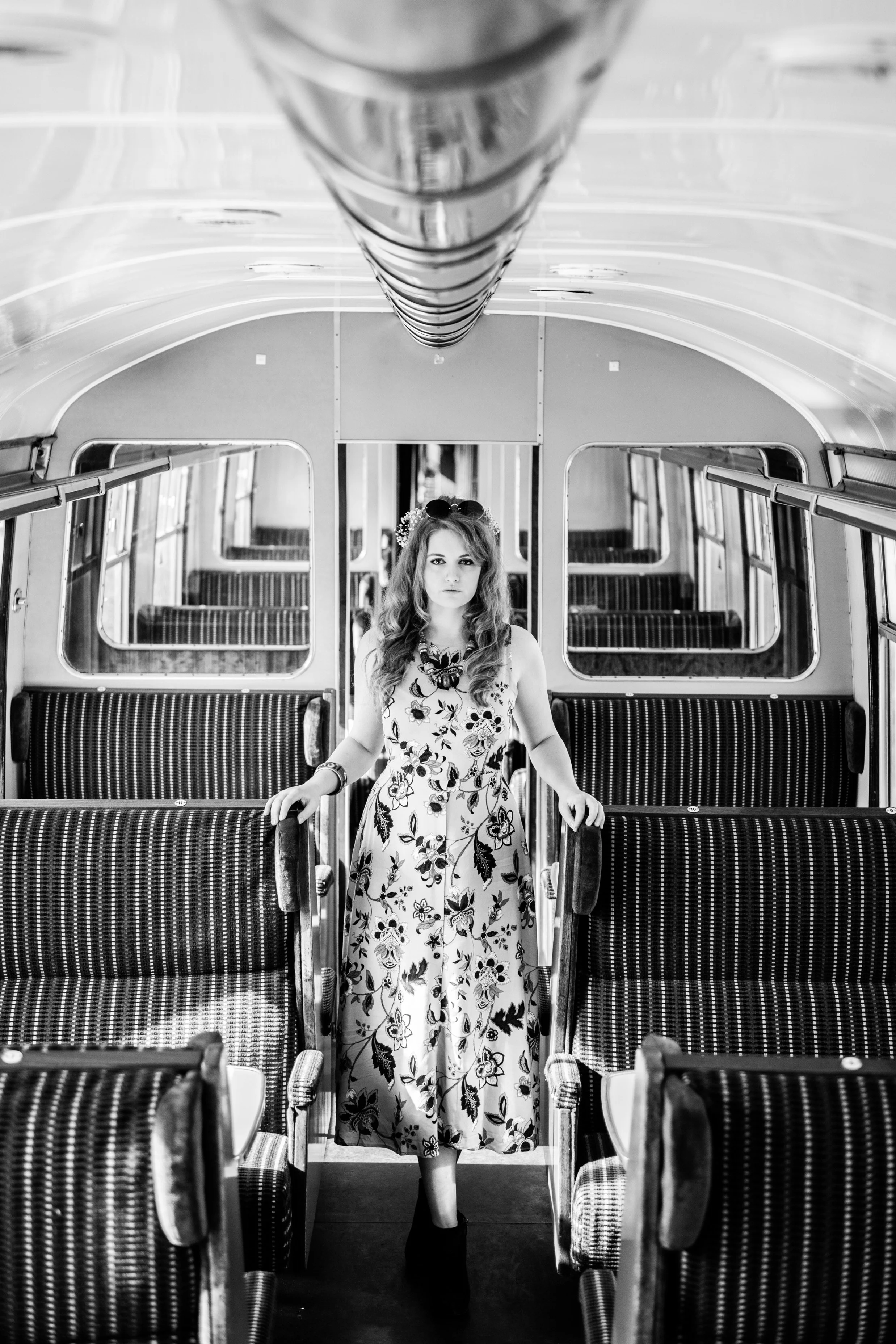 A young woman standing inside an empty vintage train car, wearing a floral dress and dark shoes, with her hair styled in loose waves.
