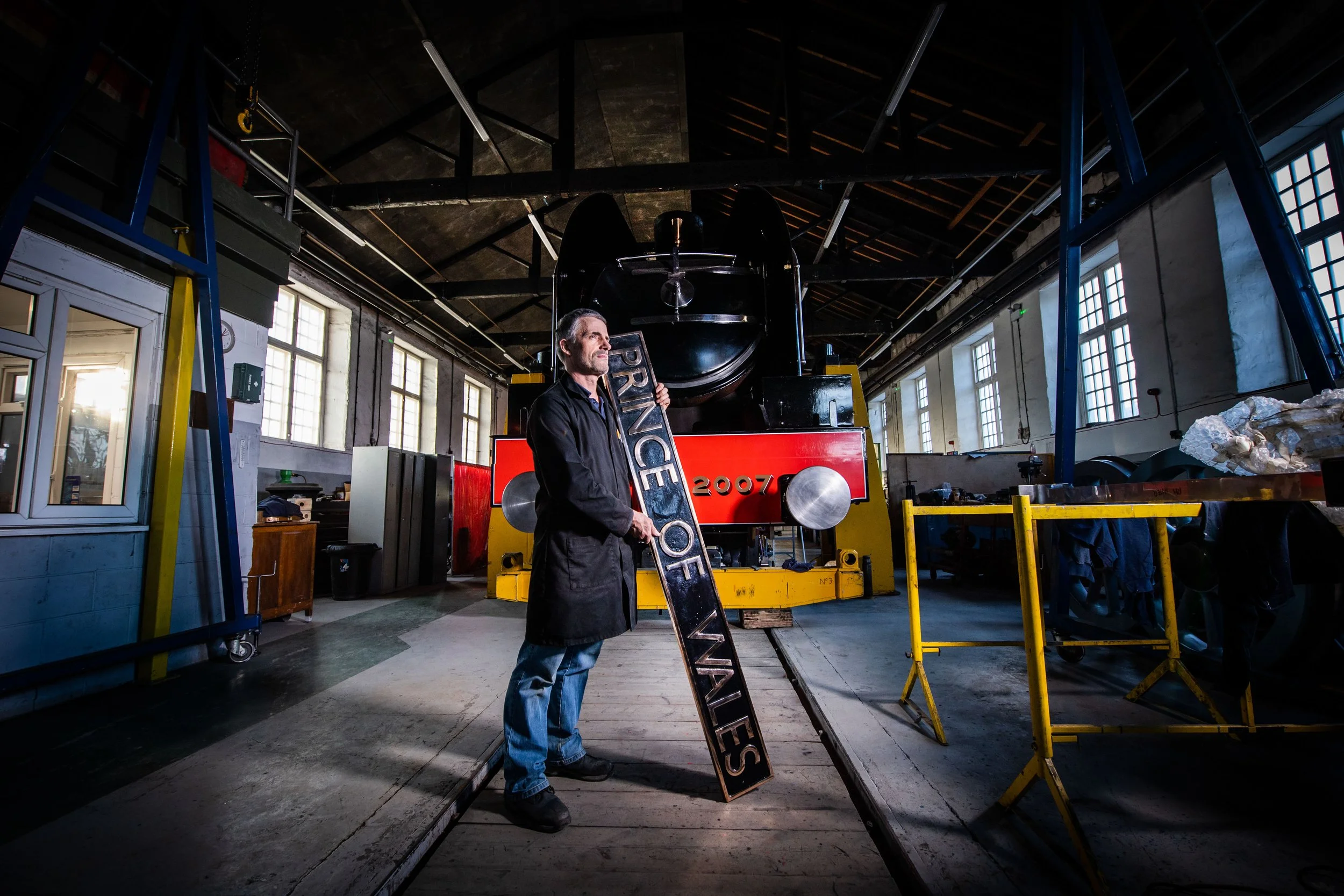 A man standing in a workshop holding a sign that reads 'PRINCE OF VALVES,' with a large vintage steam engine in the background and various equipment and tools around.