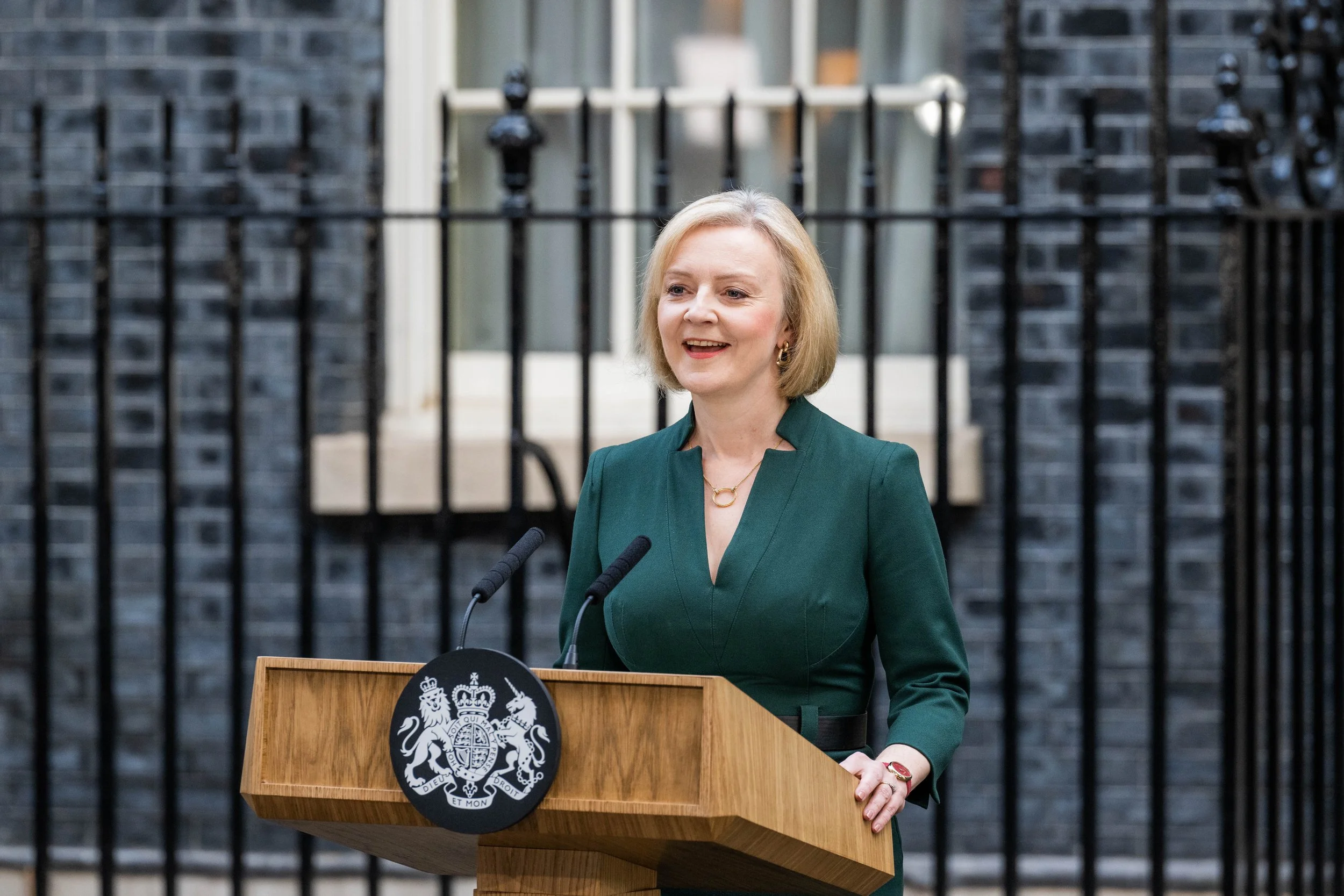 A woman with blonde hair wearing a green dress, standing at a wooden podium with a government emblem, speaking outdoors in front of a black iron fence and brick building.