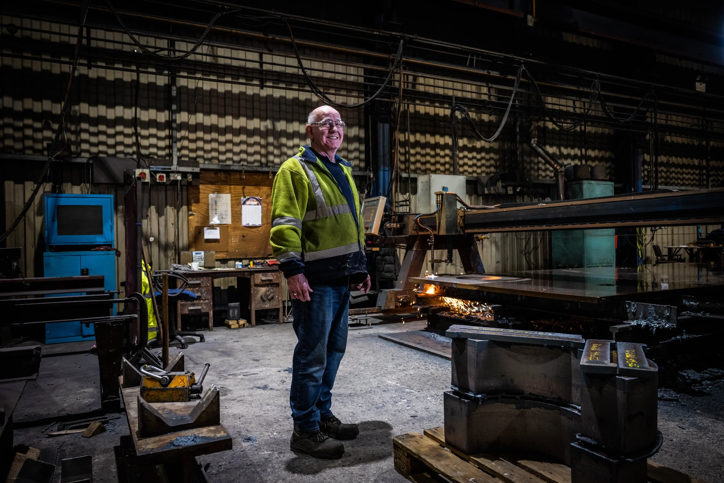 A man in safety vest and glasses standing in a metal workshop near a large industrial machine with sparks flying.