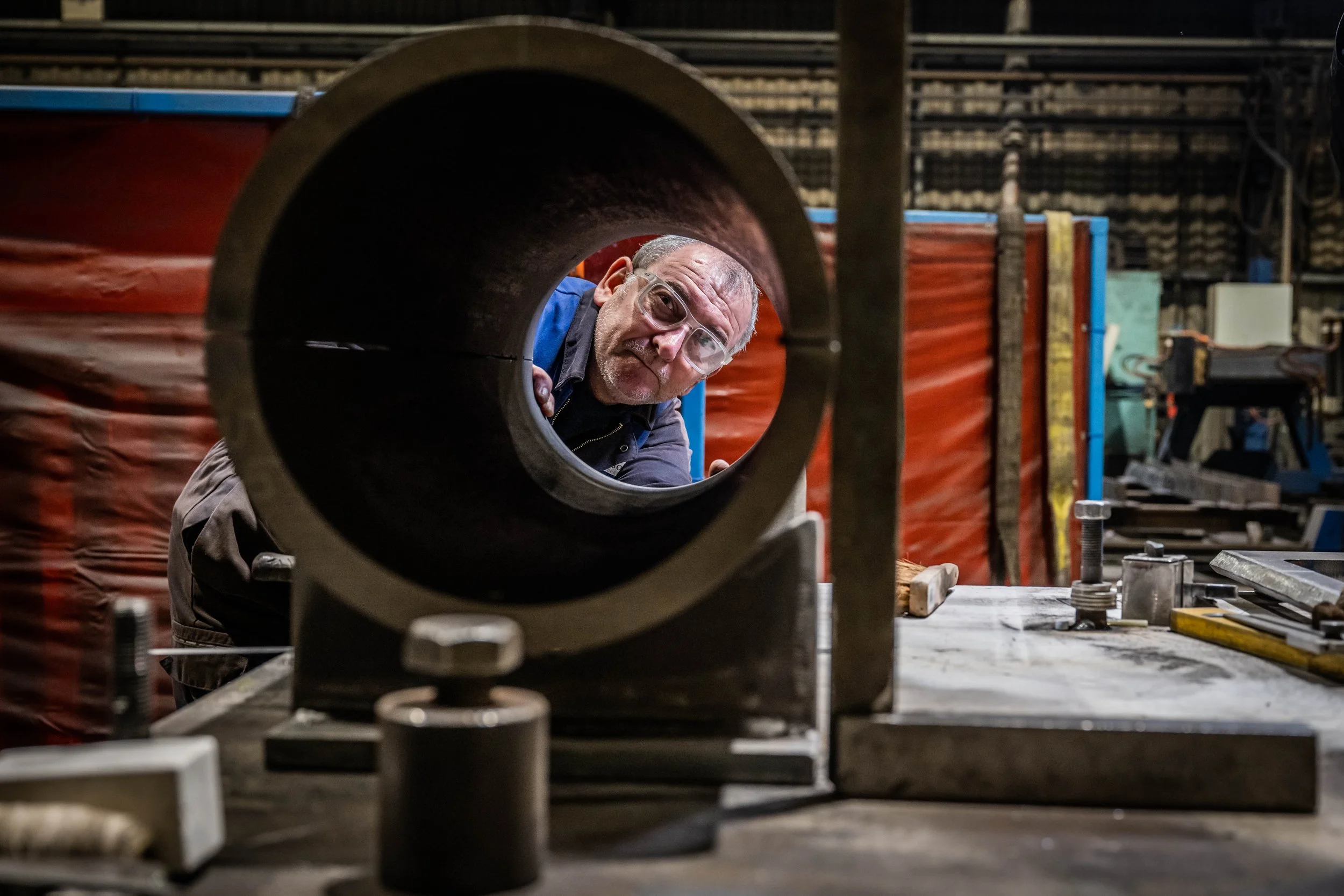 A man wearing glasses looks through a large metal pipe in a workshop.