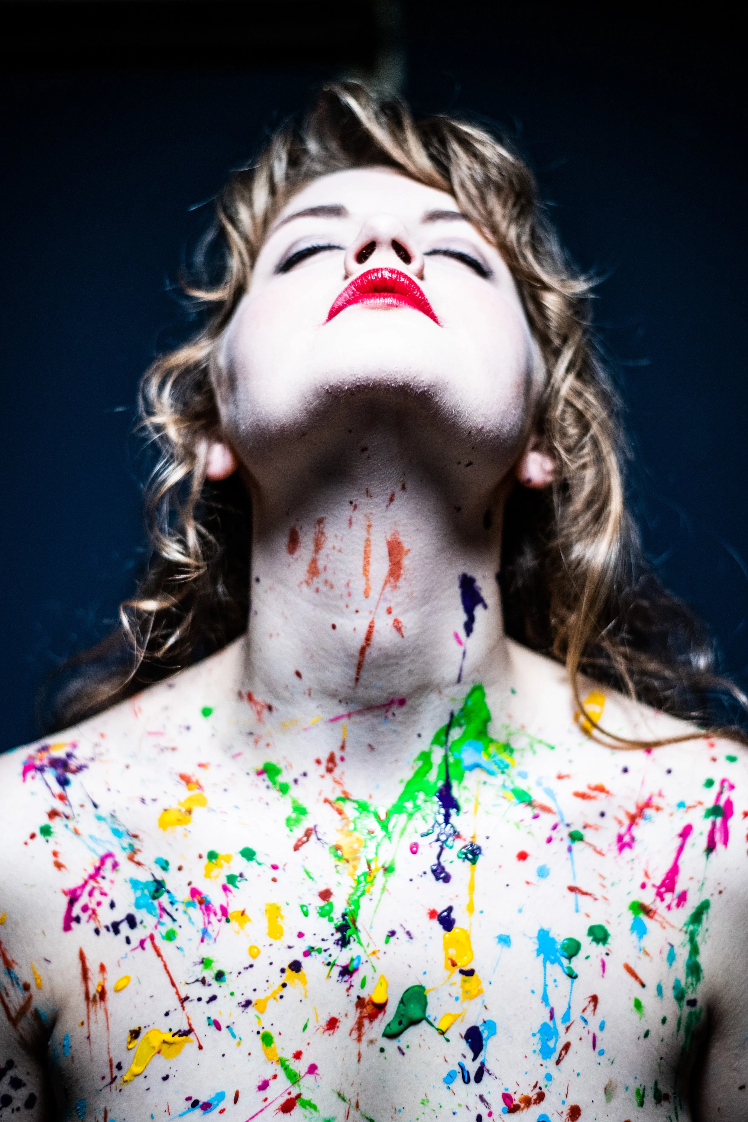 A woman with curly hair, wearing bold red lipstick, with her eyes closed and head tilted back, covered in colorful paint splatters on her neck and shoulders against a dark background.