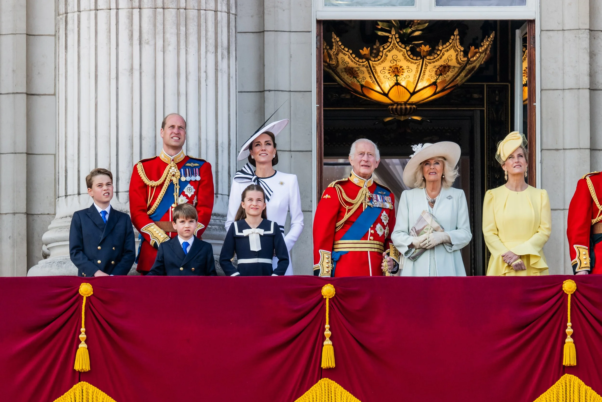 Members of the British royal family standing on a balcony during a public celebration, with some members in military uniforms and others in formal attire, behind a red velvet drape.