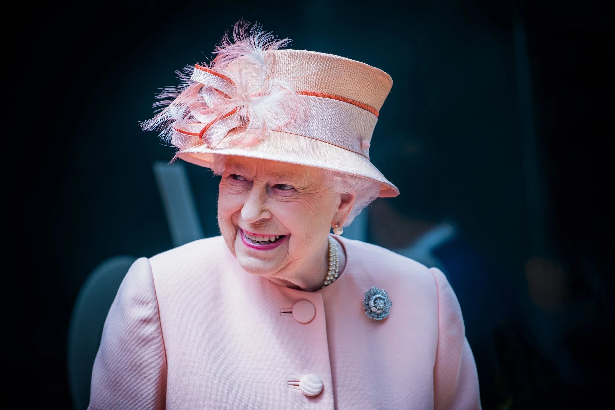 A woman wearing a pink hat with feathers, a pink coat with large buttons, a pearl necklace, and a decorative brooch, smiling and looking to the side.