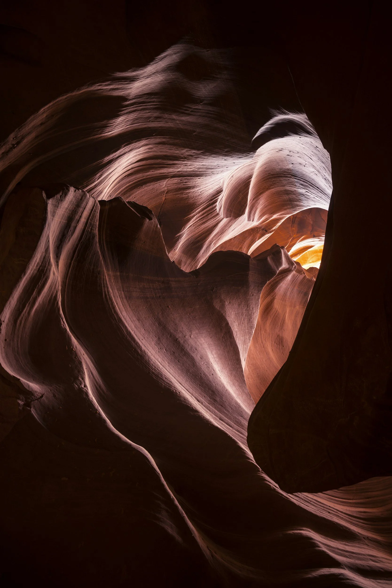 A view inside a narrow slot canyon with smooth, flowing rock formations in shades of pink, orange, and purple, illuminated by light filtering from above.