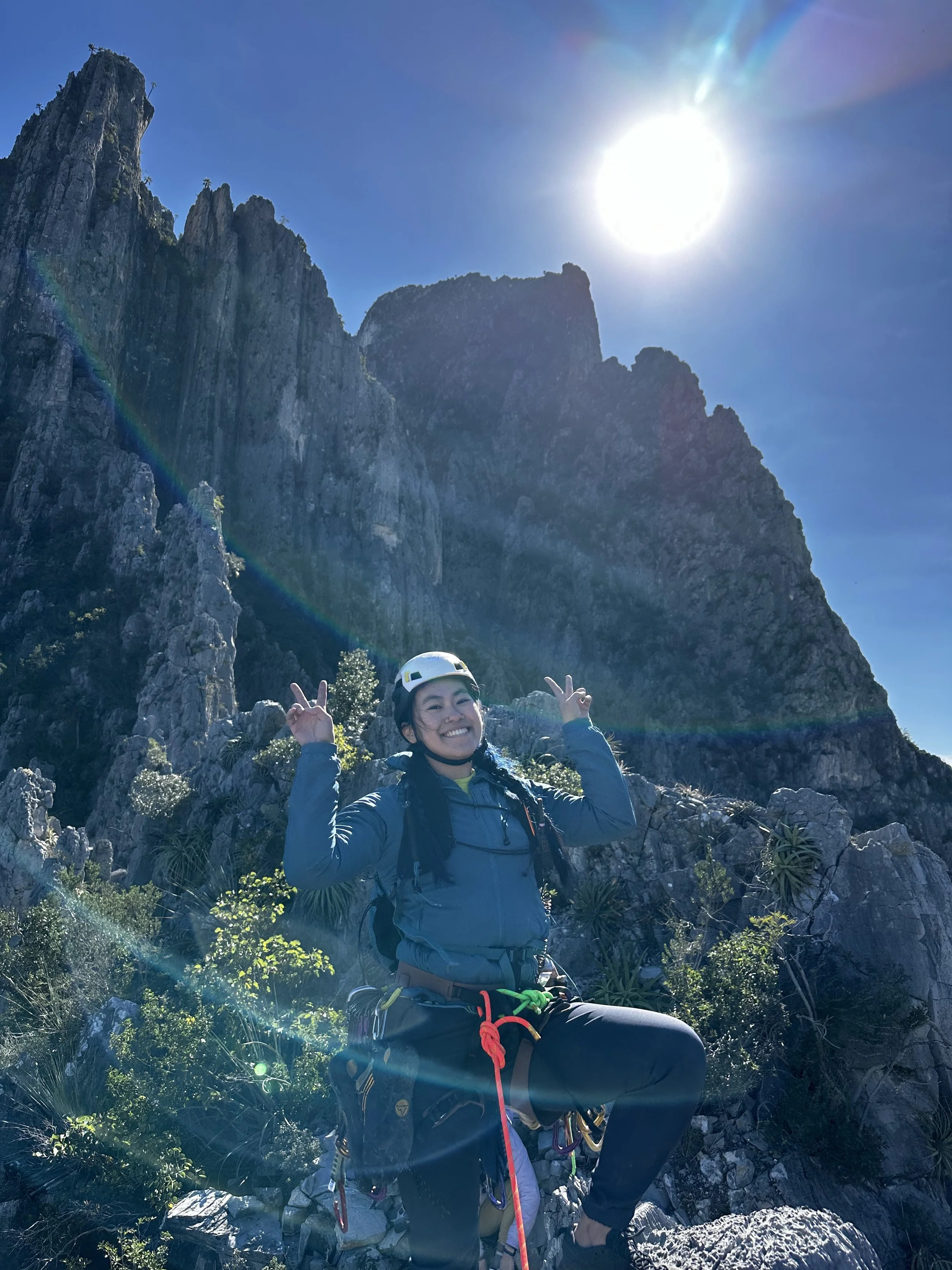 A woman climber wearing a helmet smiling and making peace signs in a mountain landscape under a bright sun, with rocky cliffs and green bushes around.