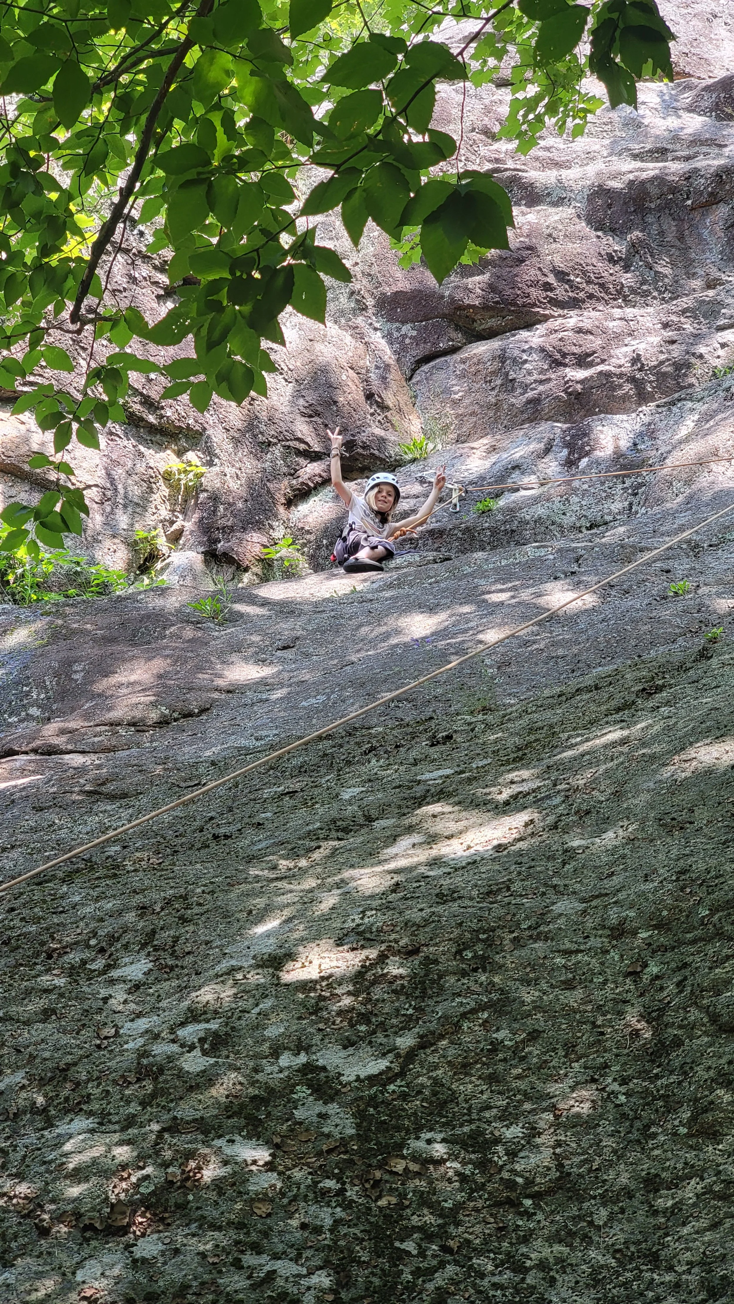 A young girl rock climbing on a large sloped rock face in a forested area, wearing a helmet and safety gear, and making a peace sign with her right hand.