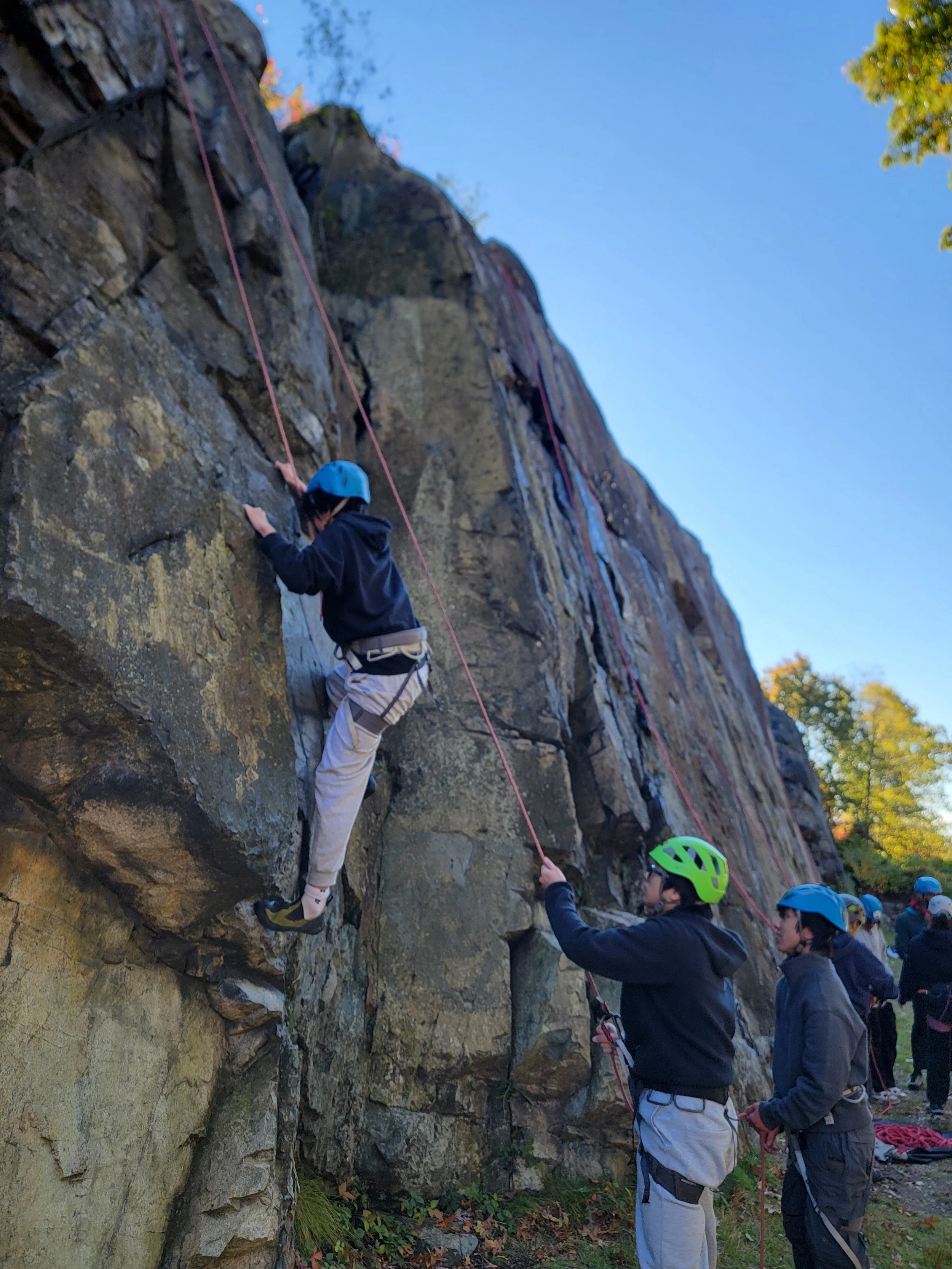 A group of children wearing helmets rock climbing on an outdoor rock wall, with one child actively climbing and others managing safety ropes.
