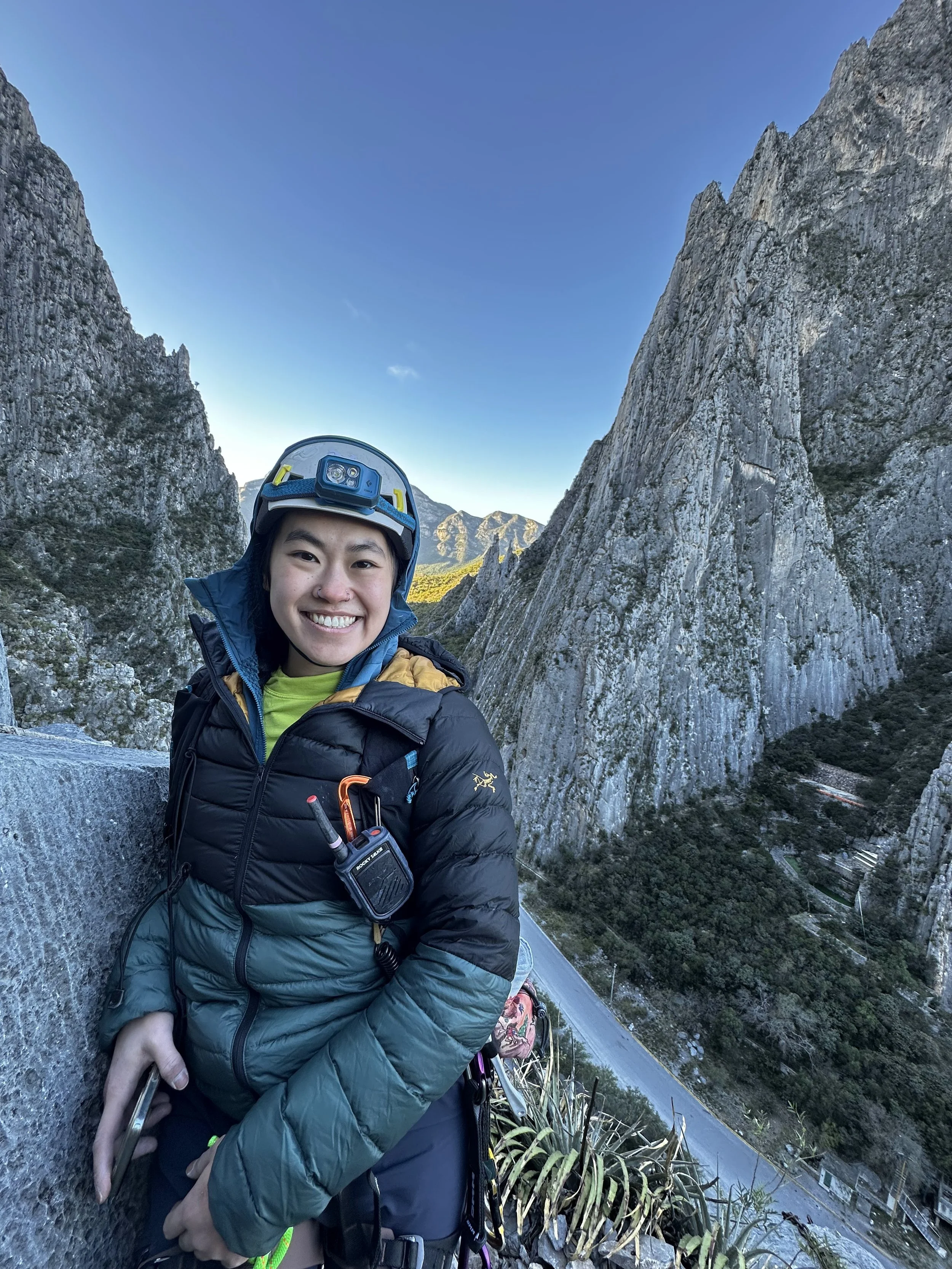 Smiling woman in climbing gear and helmet standing outdoors among large rocky mountains with a winding road below and distant mountain peaks under a clear blue sky.