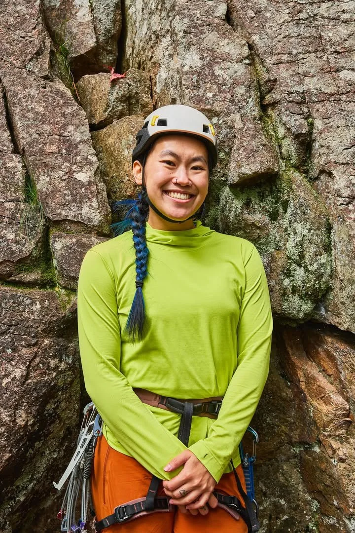 A woman in climbing gear standing in front of a rocky wall, smiling, wearing a helmet, bright green long-sleeve shirt, and orange pants.
