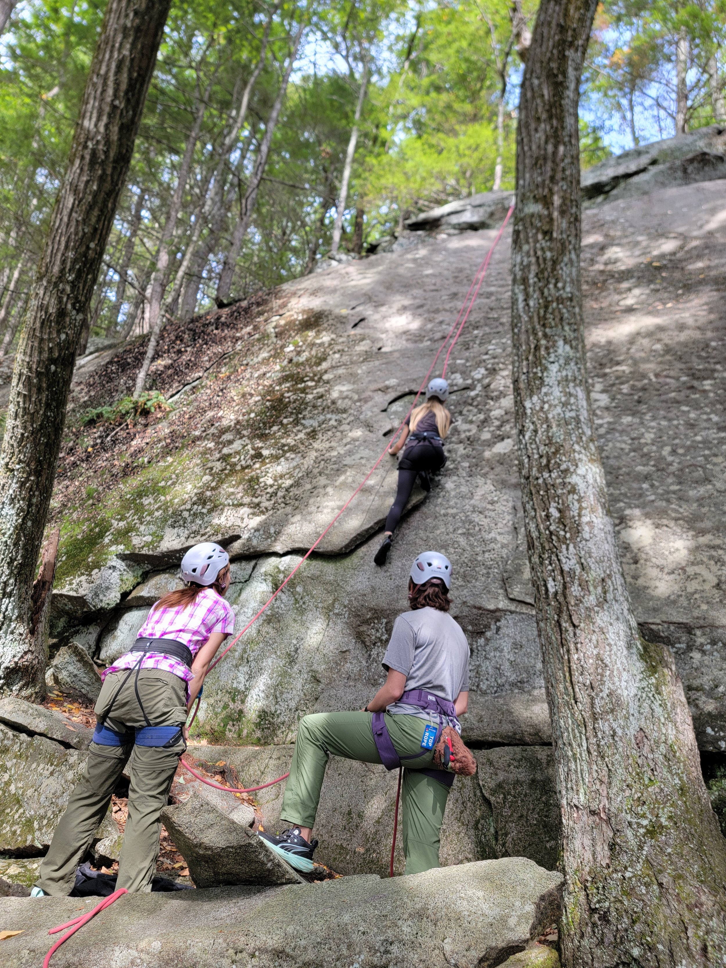 People rock climbing on a steep outdoor rock face, with two individuals spotting and one climbing.