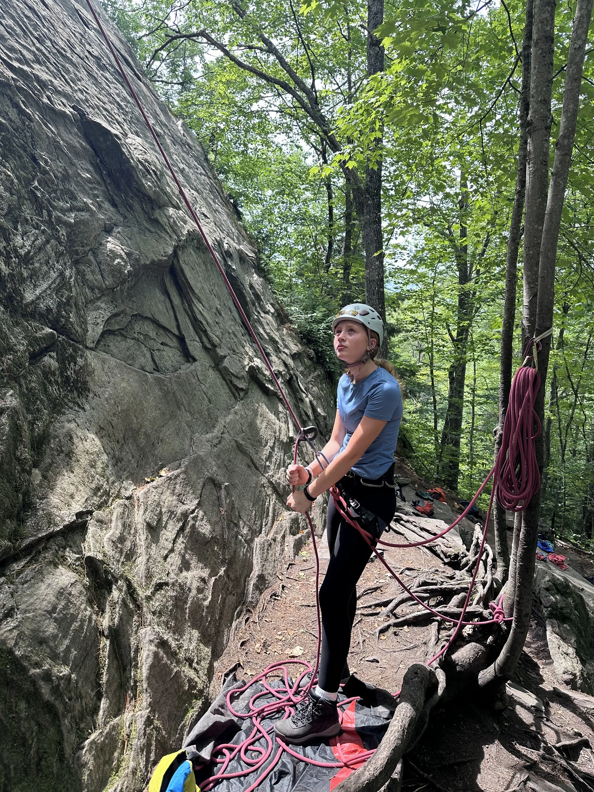 A young woman wearing a helmet and climbing gear standing on a rocky trail in a forest, preparing to climb a rock wall.