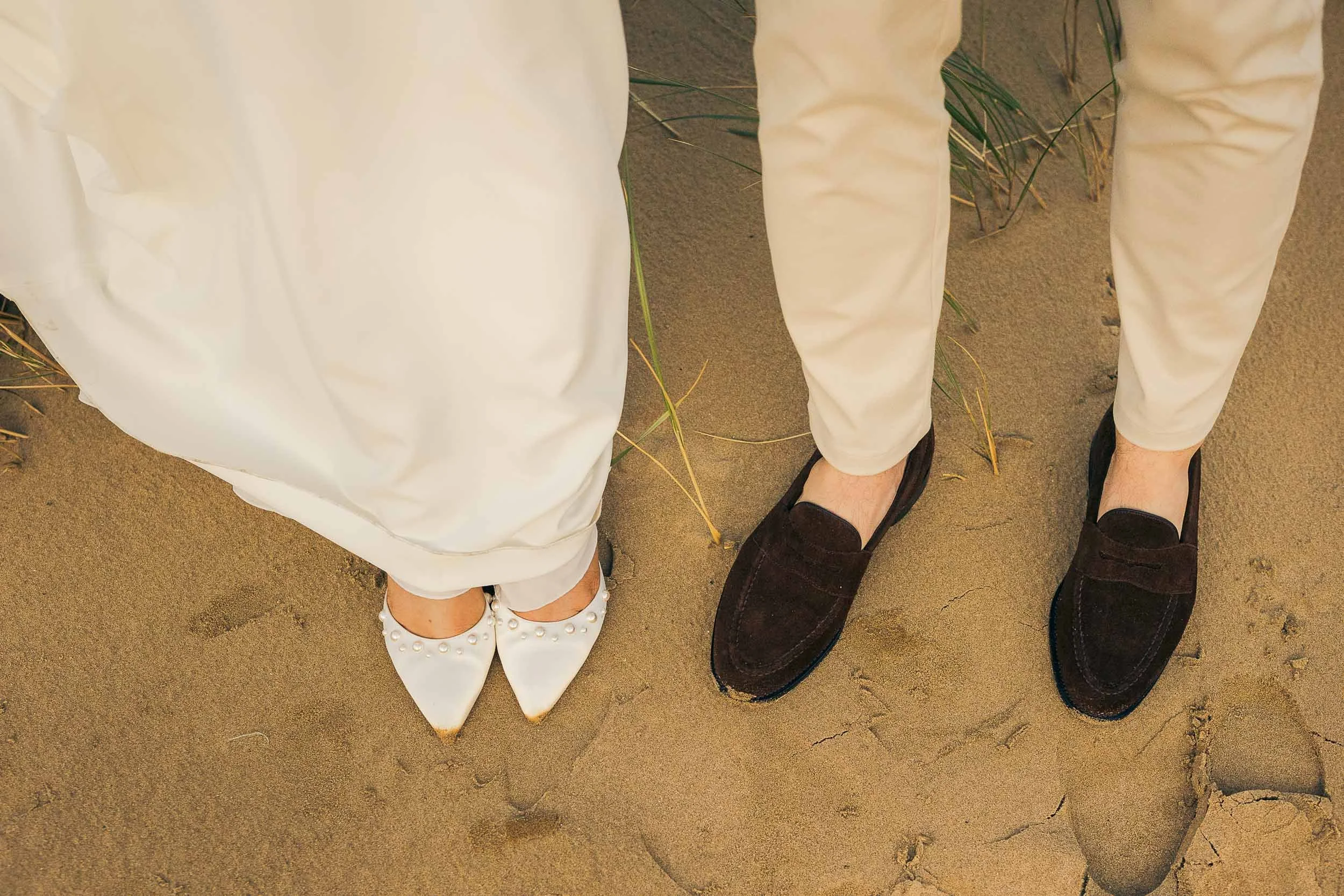 Close-up of a woman wearing white shoes with pearls and a man wearing brown loafers, standing on sand with some beach grass.